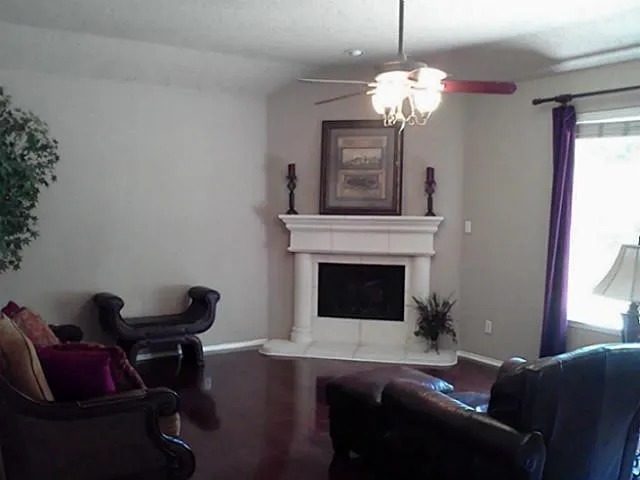 Corner fireplace with a mantel, dark wood-finish flooring, and a ceiling fan with integrated lighting