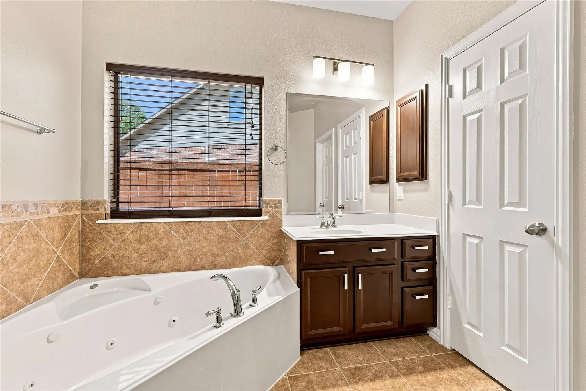 Jetted soaking tub with chrome fixtures, wood-finish vanity with white countertop, and tiled flooring
