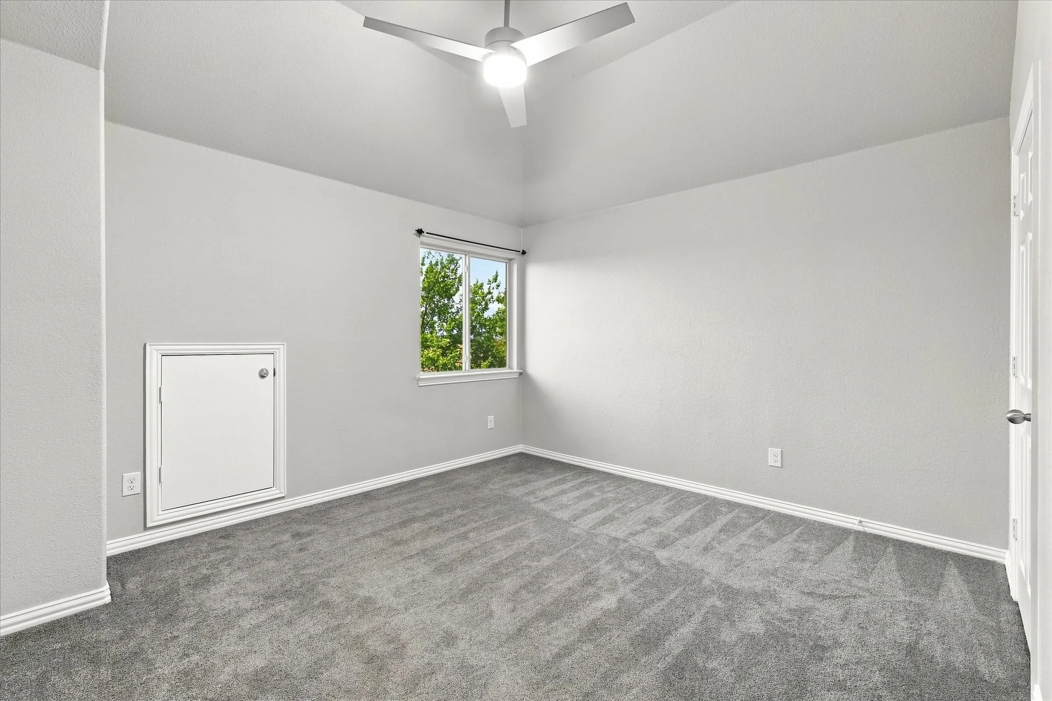 Vaulted ceiling with a ceiling fan, light gray wall paint, gray carpet flooring, window with exterior landscaping, and white trim