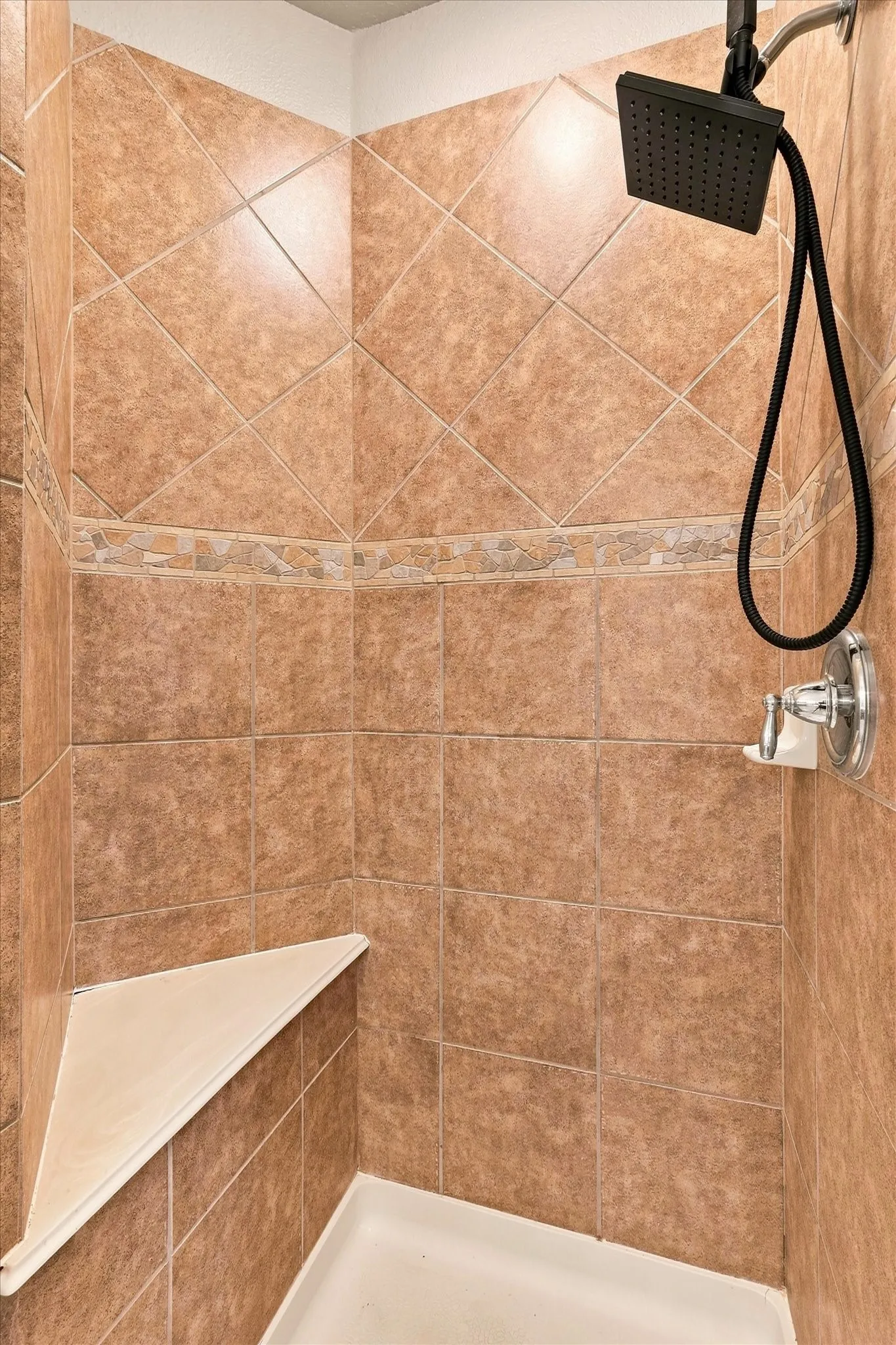 Shower stall with brown tile walls, decorative stone accent band, corner bench, and black square shower head with handheld attachment