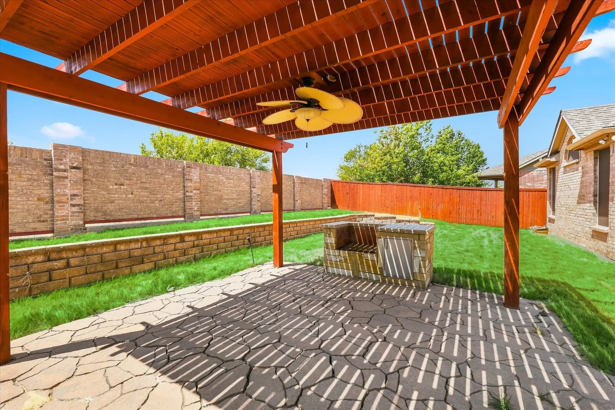 Expansive backyard featuring a wood pergola with a ceiling fan, a stone patio, a built-in stone grill station, a manicured lawn, and brick perimeter fencing