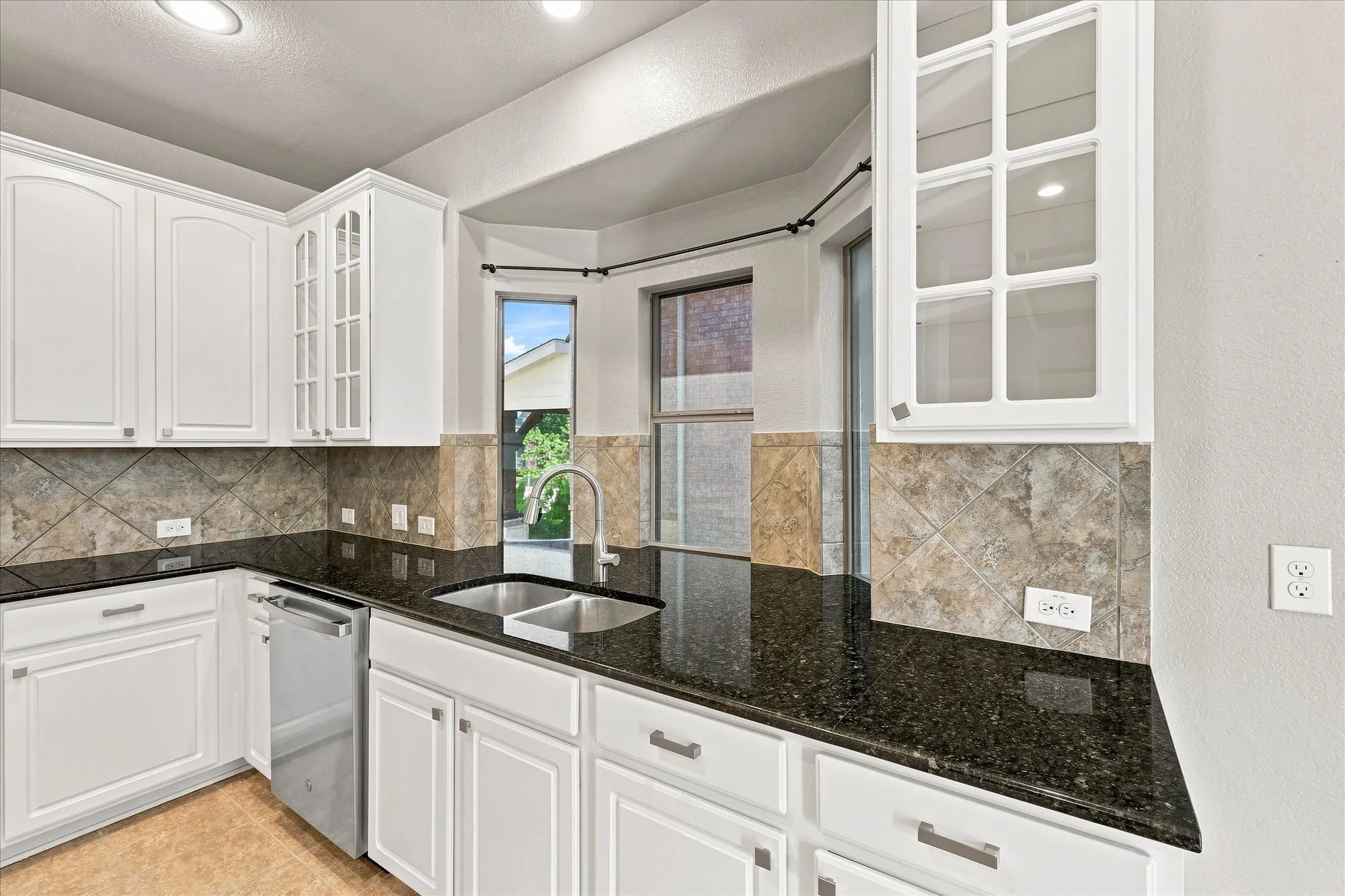White cabinetry, dark stone countertops, tile backsplash, stainless steel sink, and built-in dishwasher
