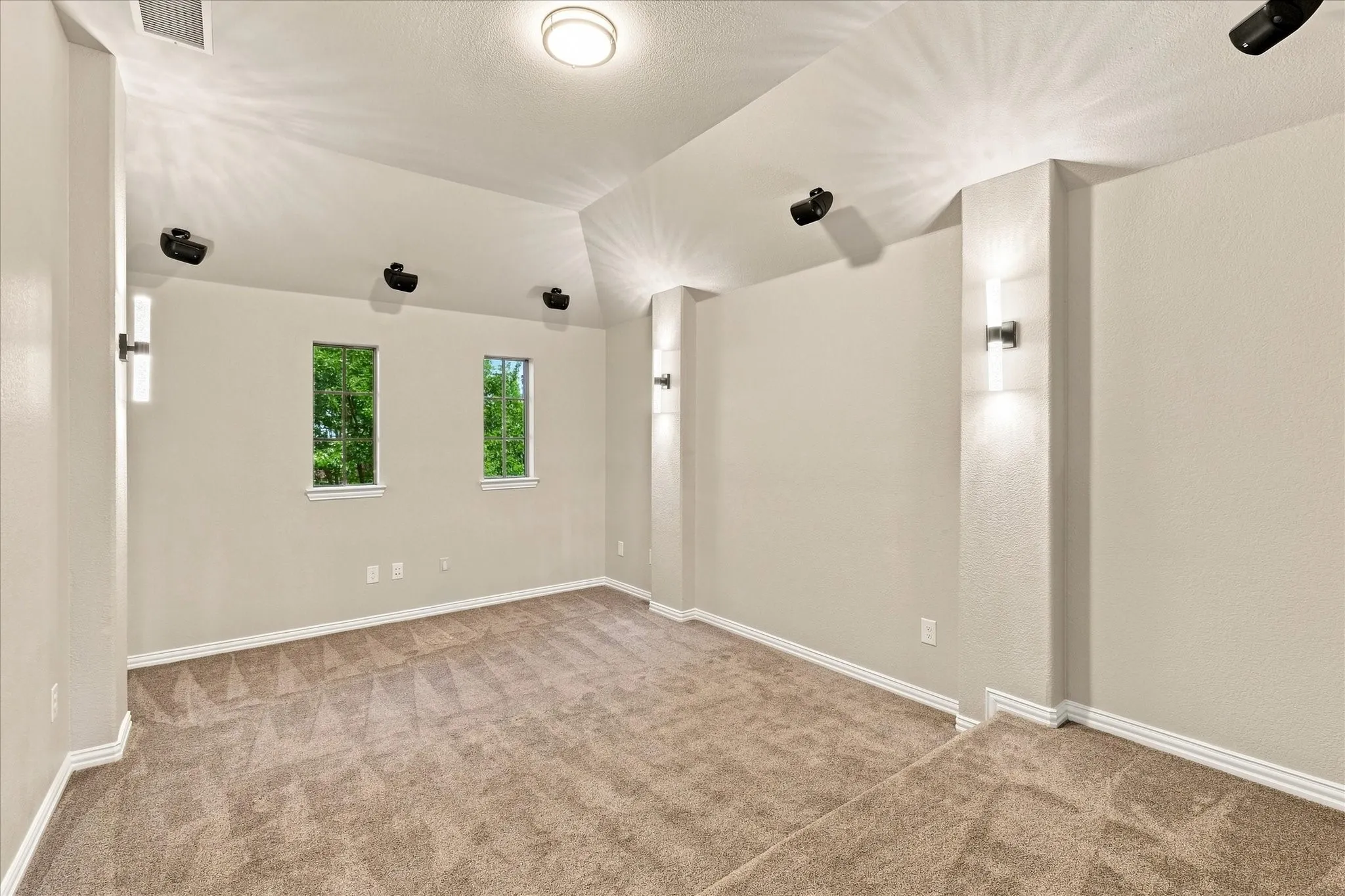 Recessed ceiling speakers, dual wall sconces, ceiling-mounted light fixture, two vertical windows, and plush carpeting