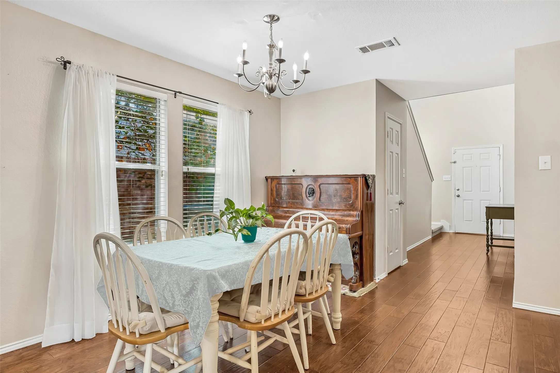 Wood-finish flooring throughout, dual windows with blinds, a polished chandelier, and a white paneled entry door
