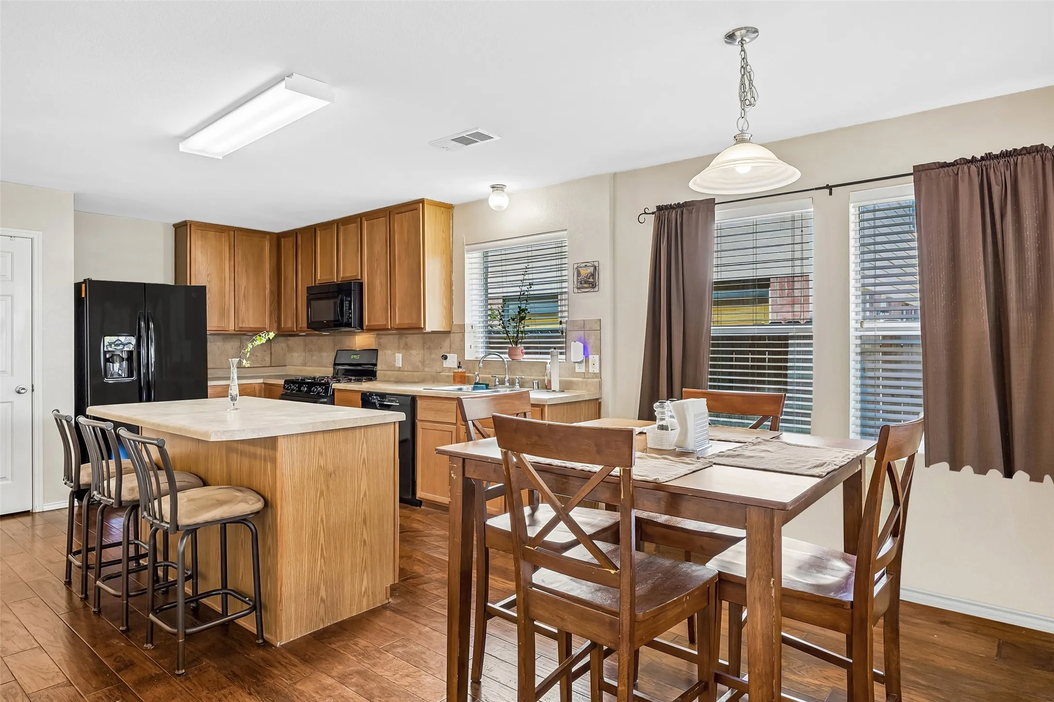 Kitchen featuring wood-finish flooring, a central island with countertop seating, wood cabinetry, a tile backsplash, and black appliances