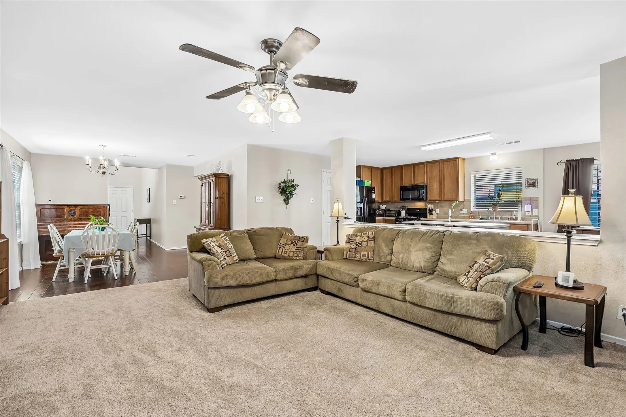 Expansive living area with neutral carpeting, an overhead ceiling fan with integrated lighting, and direct access to a kitchen featuring wood cabinetry and black appliances