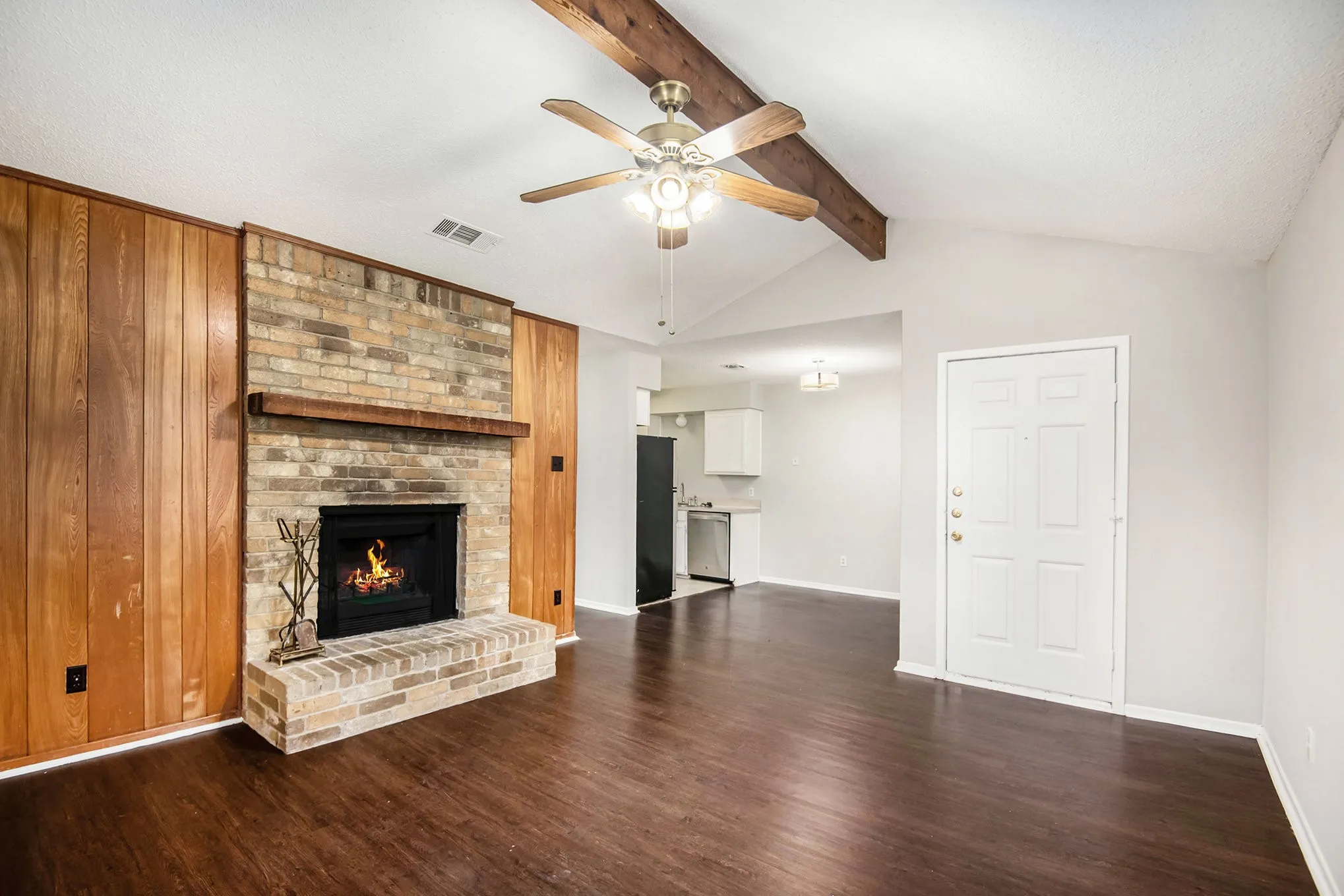 Brick fireplace with a wood mantle, wood-finish flooring, vaulted ceiling with exposed beam, and a ceiling fan