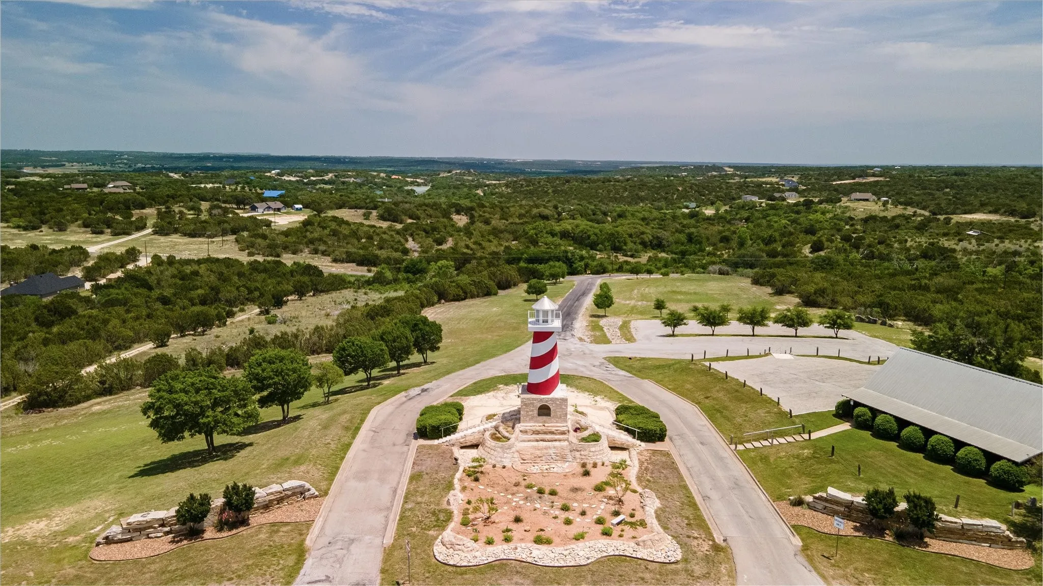 Red and white striped lighthouse structure, stone base, paved driveways, surrounding green landscaping, and distant wooded terrain
