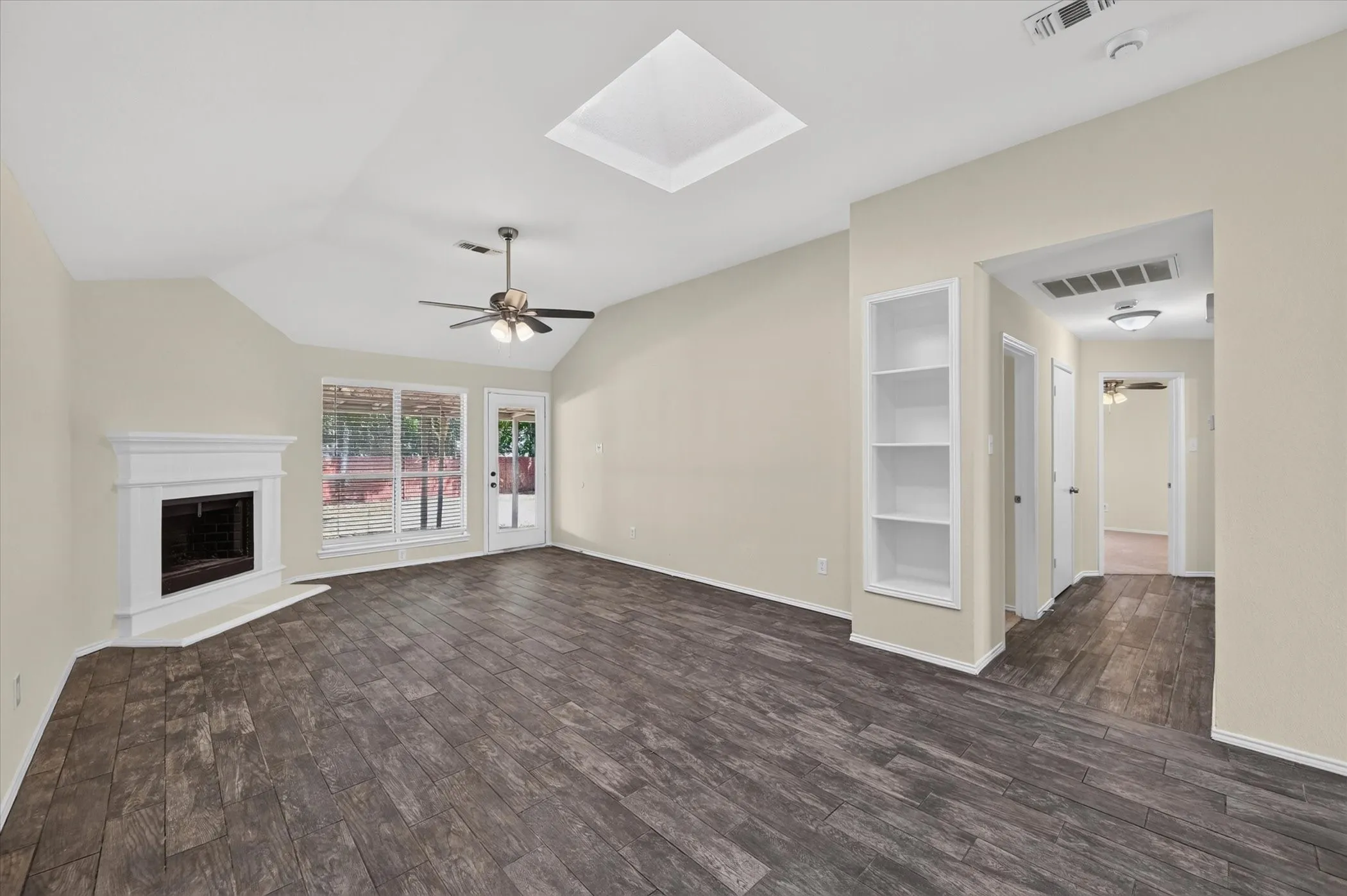 Vaulted ceiling with skylight, wood-finish flooring, white mantel fireplace, ceiling fan, and built-in shelving