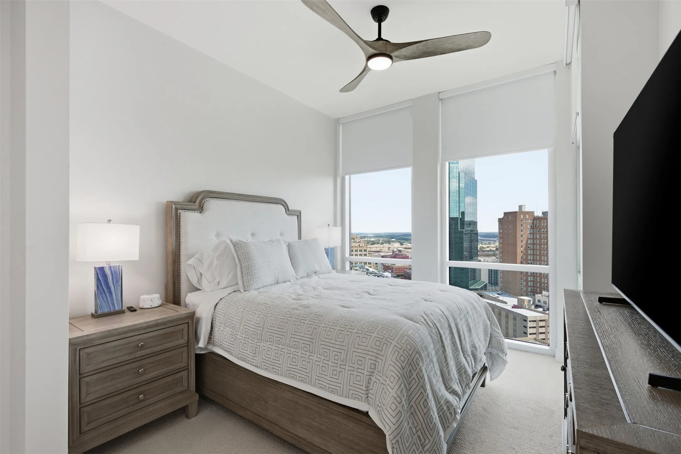 Expansive windows with roller shades, light-colored carpeting, and a ceiling fan