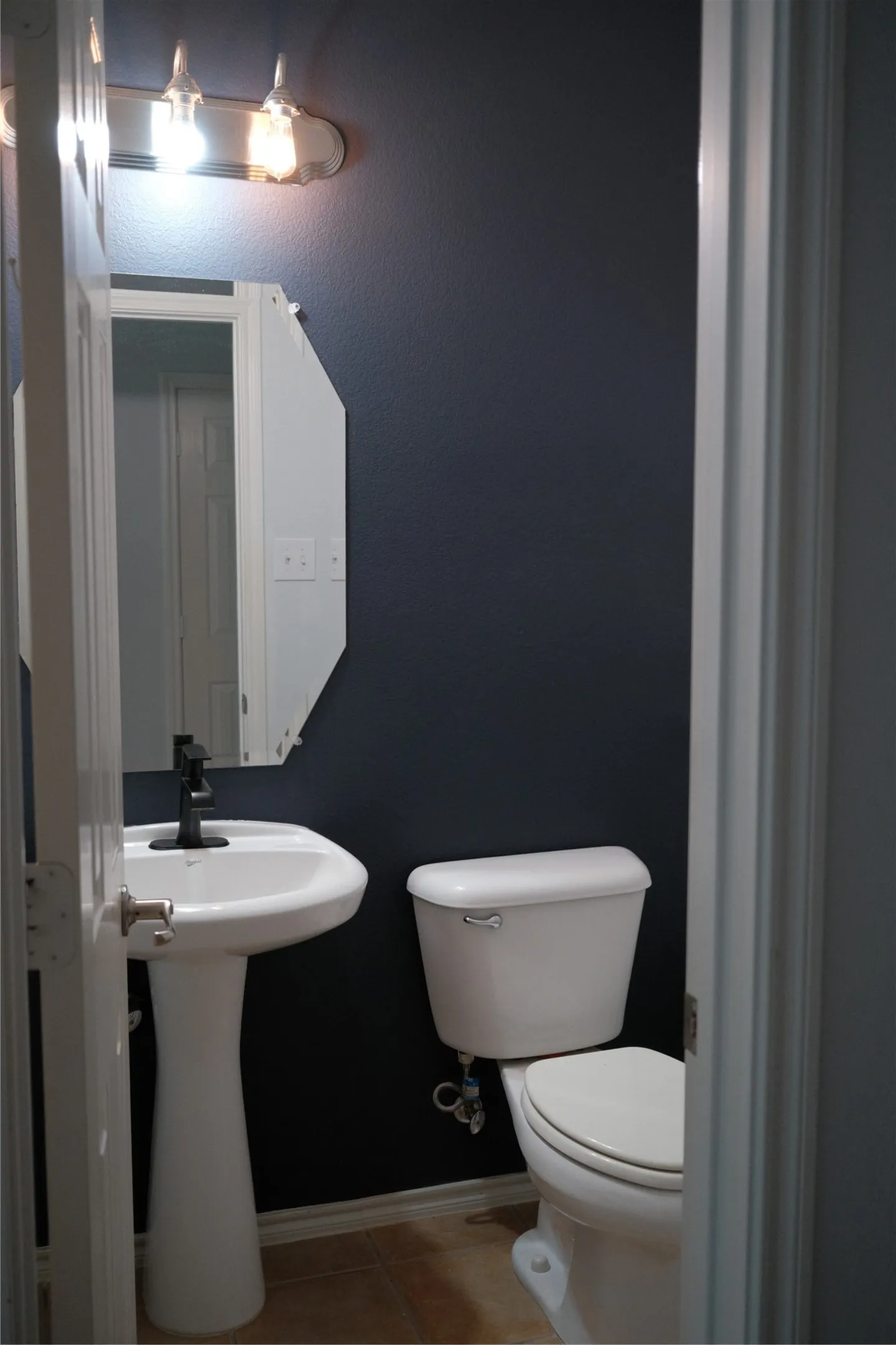 Compact bathroom featuring a pedestal sink with chrome fixtures, an elongated toilet, wood-finish flooring, and dark accent walls