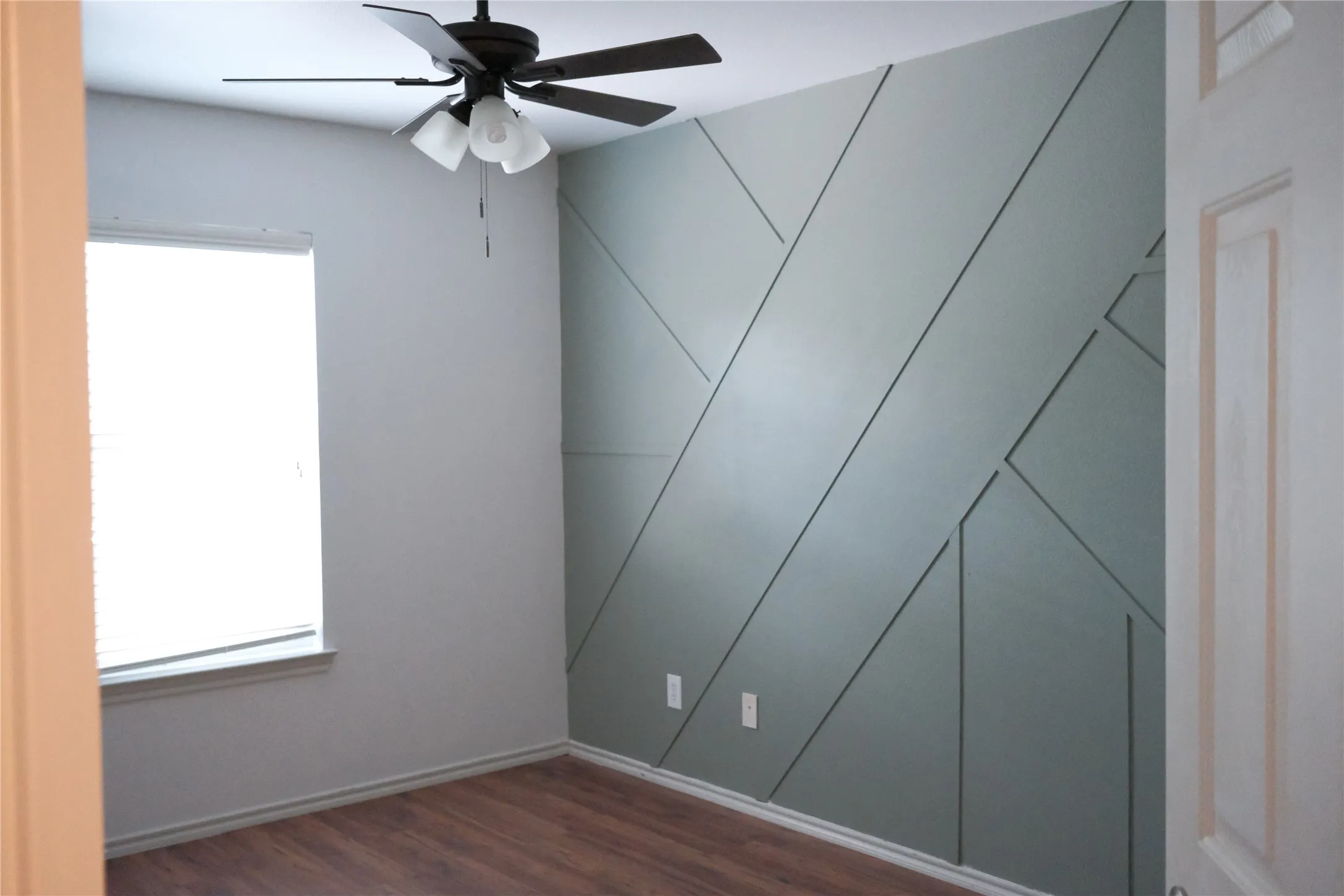 Geometric accent wall, wood-finish flooring, ceiling fan with integrated lighting, and window with blinds