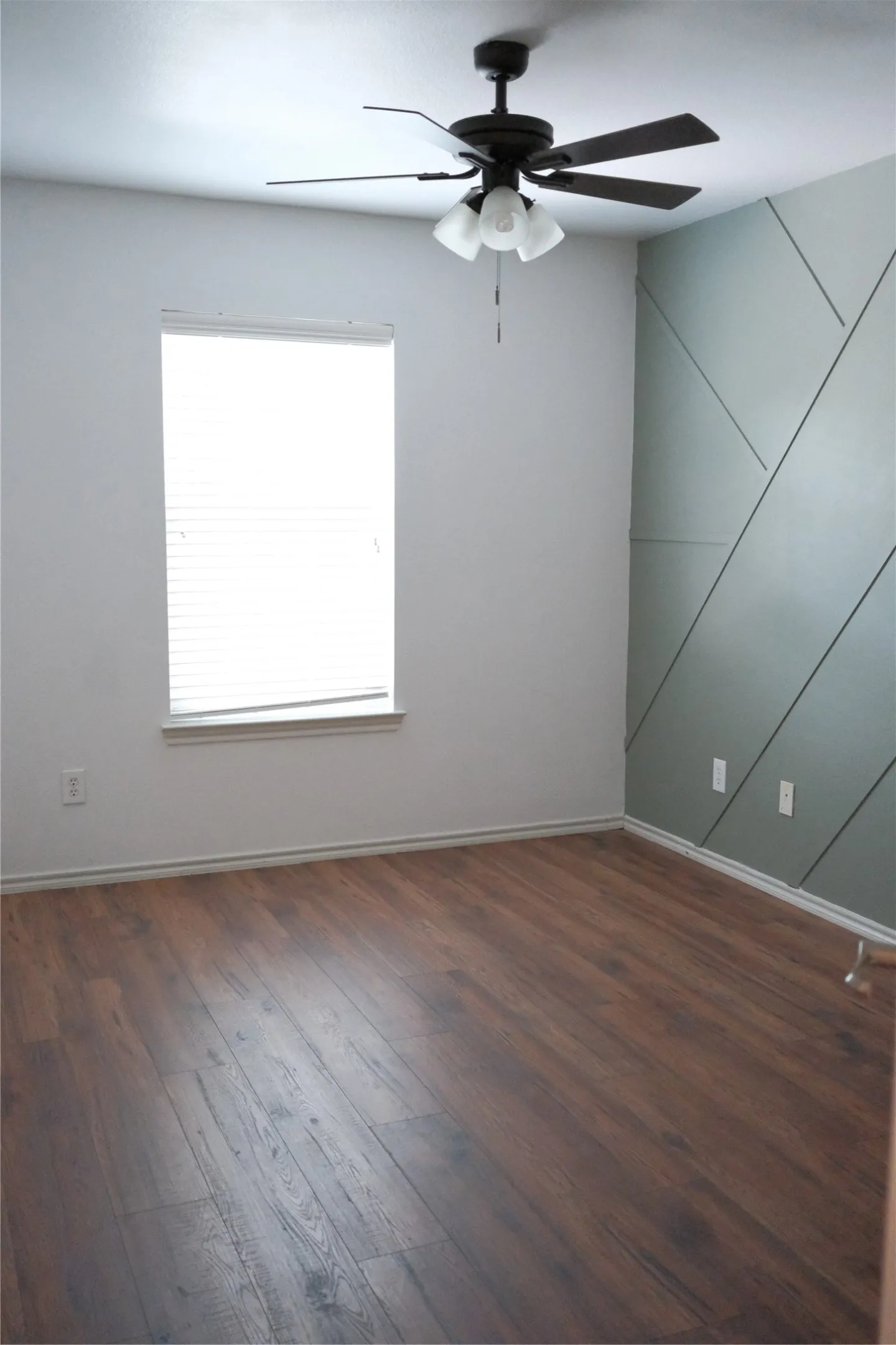 Wood-finish flooring, accent wall with geometric trim, single window with blinds, and a ceiling fan with integrated lighting