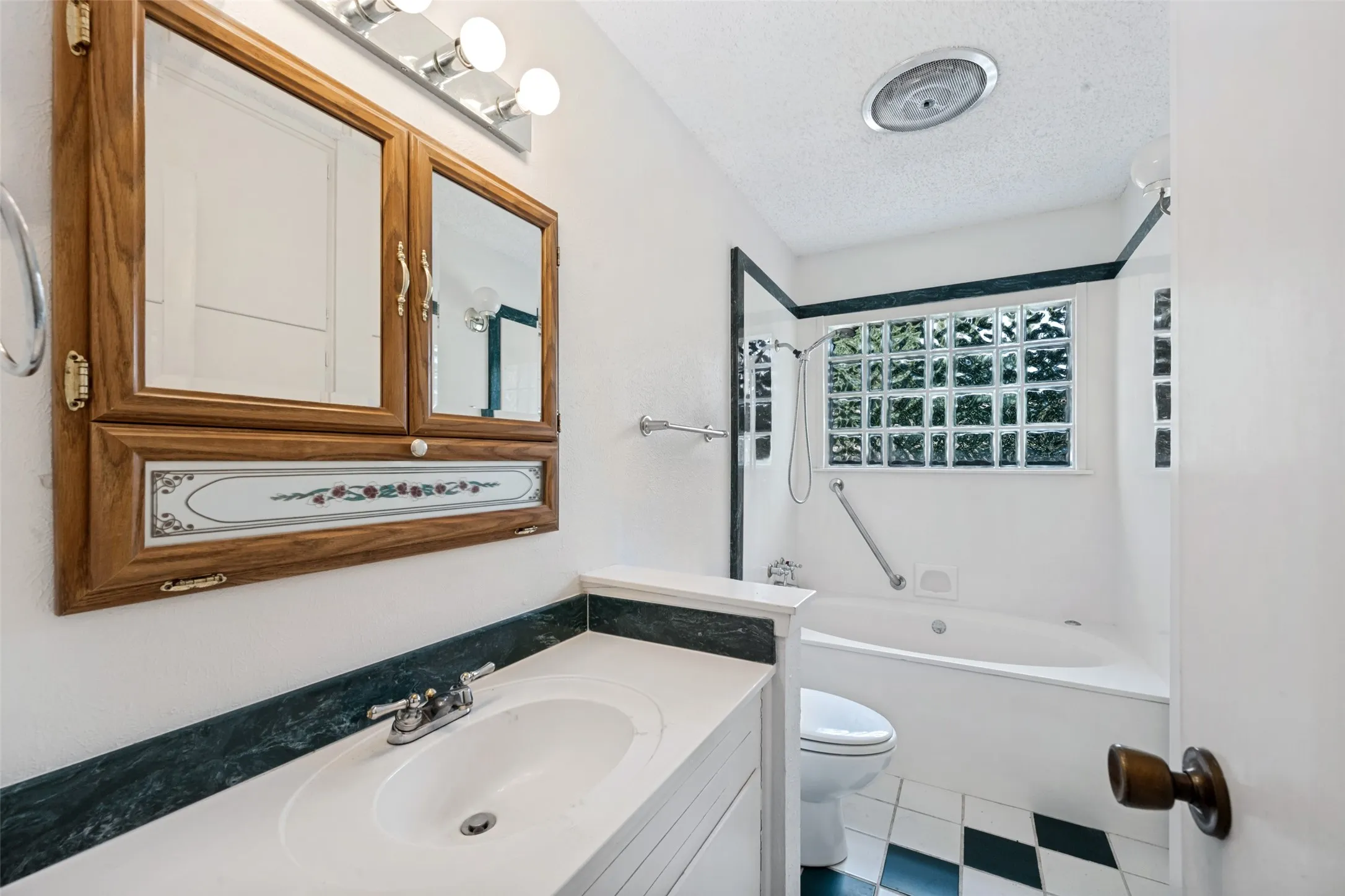 Bathroom featuring a built-in tub, glass block window, wood-finish medicine cabinet, vanity with integrated sink, and checkered floor tile