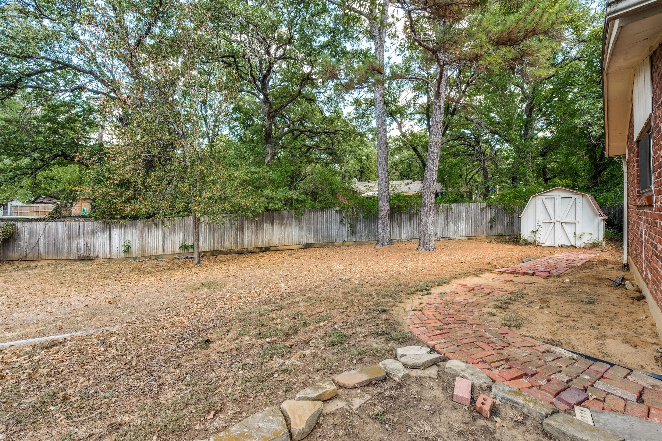Expansive backyard featuring mature trees, a wood privacy fence, a brick paver pathway, and a white storage shed