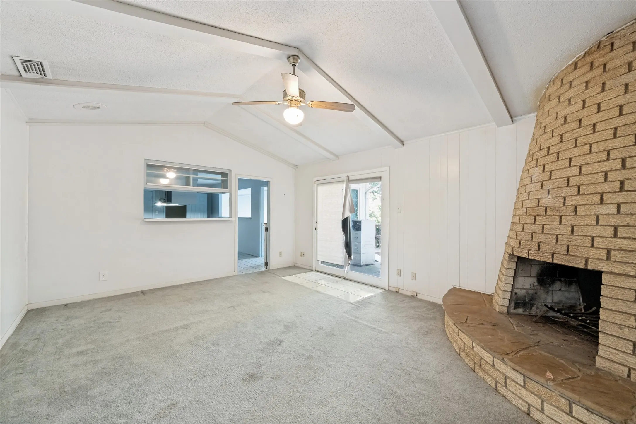 Vaulted ceiling with exposed beams, brick fireplace with stone hearth, pass-through window, ceiling fan, and sliding glass door access