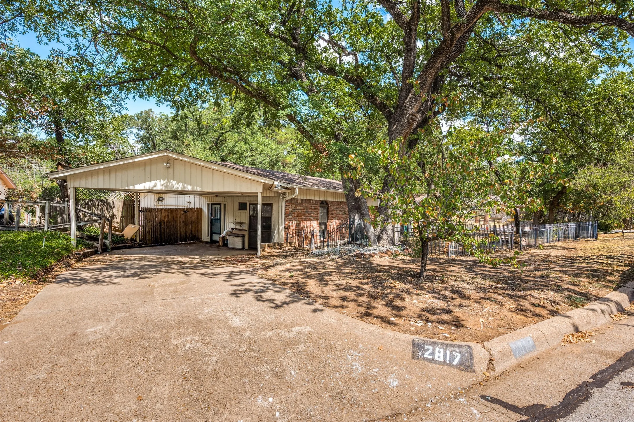 Brick exterior with a covered carport, mature trees, and a concrete driveway