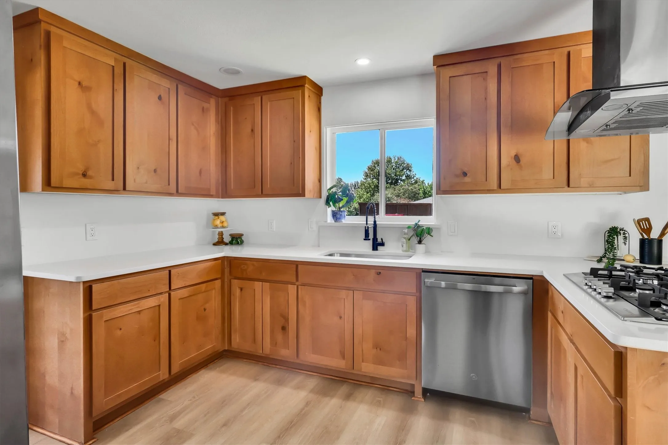 Wood-finish flooring, wood cabinetry, built-in stainless steel dishwasher, stainless steel gas range with coordinating hood, and white countertops