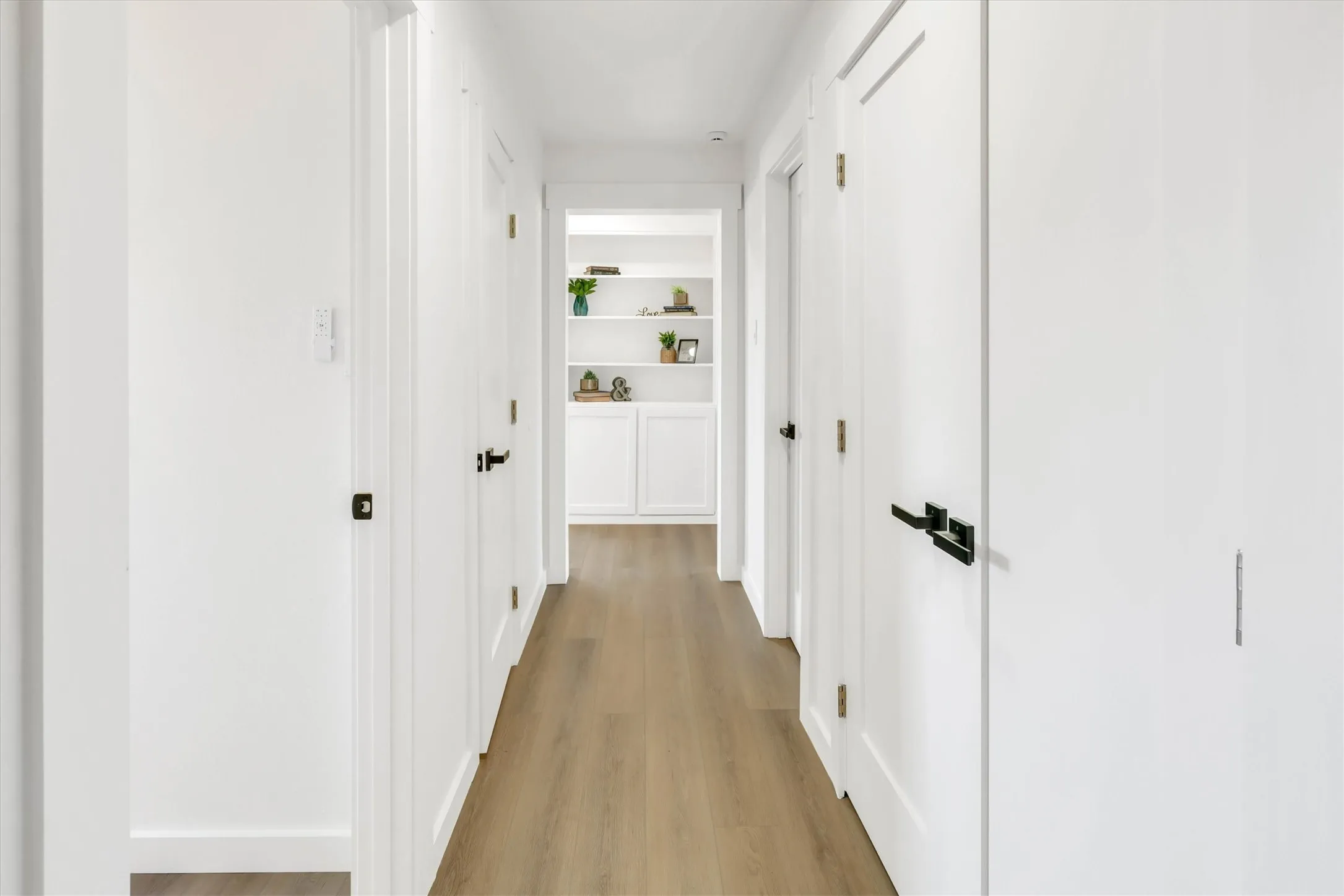 Hallway featuring wood-finish flooring, white walls, white doors with dark hardware, and built-in shelving