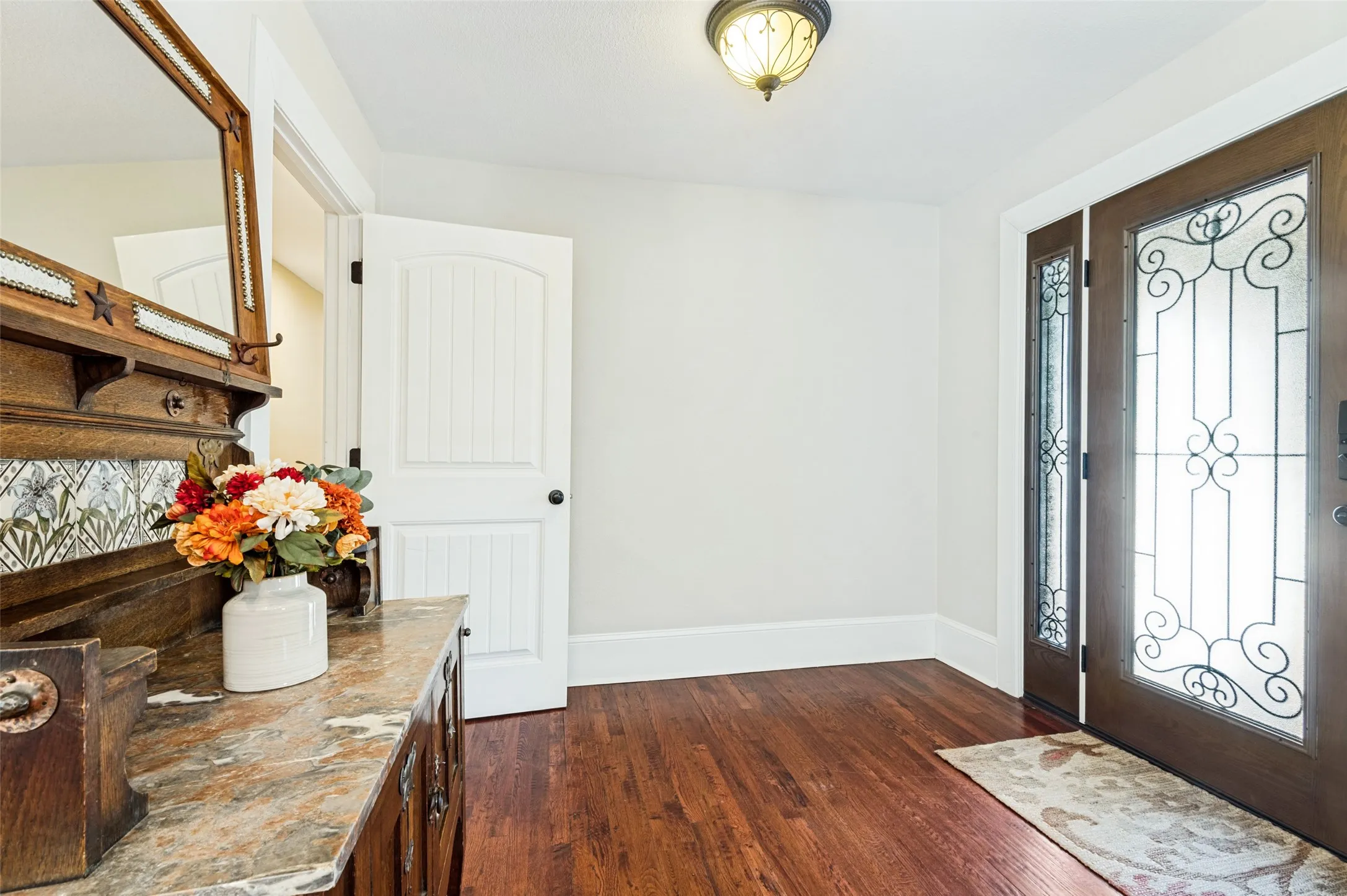 Rich wood-finish flooring, solid core entry door with decorative ironwork and frosted glass, recessed side window, and ceiling-mounted light fixture