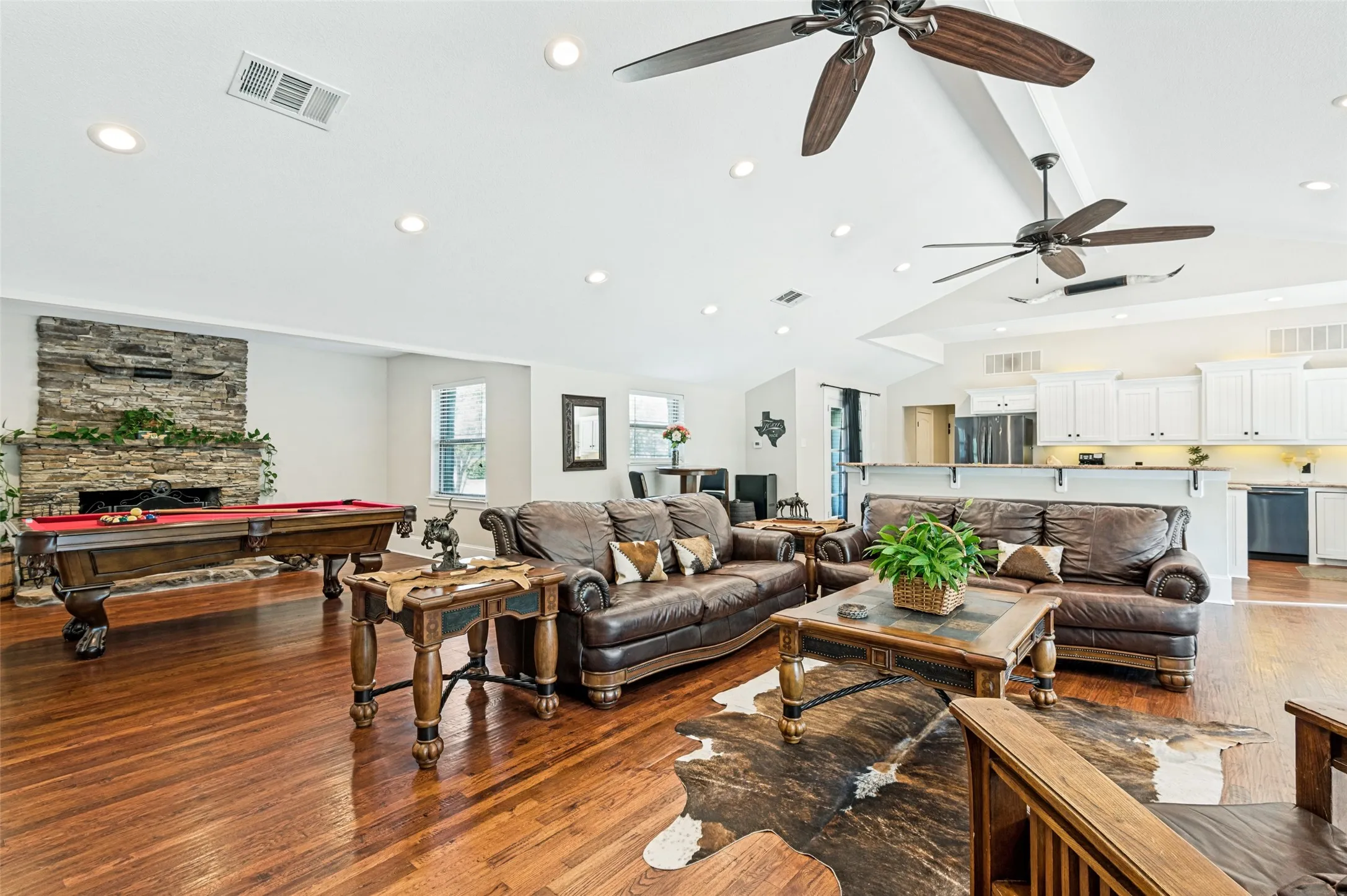 Open-concept living area featuring vaulted ceilings, wood-finish flooring, a stacked stone fireplace, recessed lighting, and ceiling fans