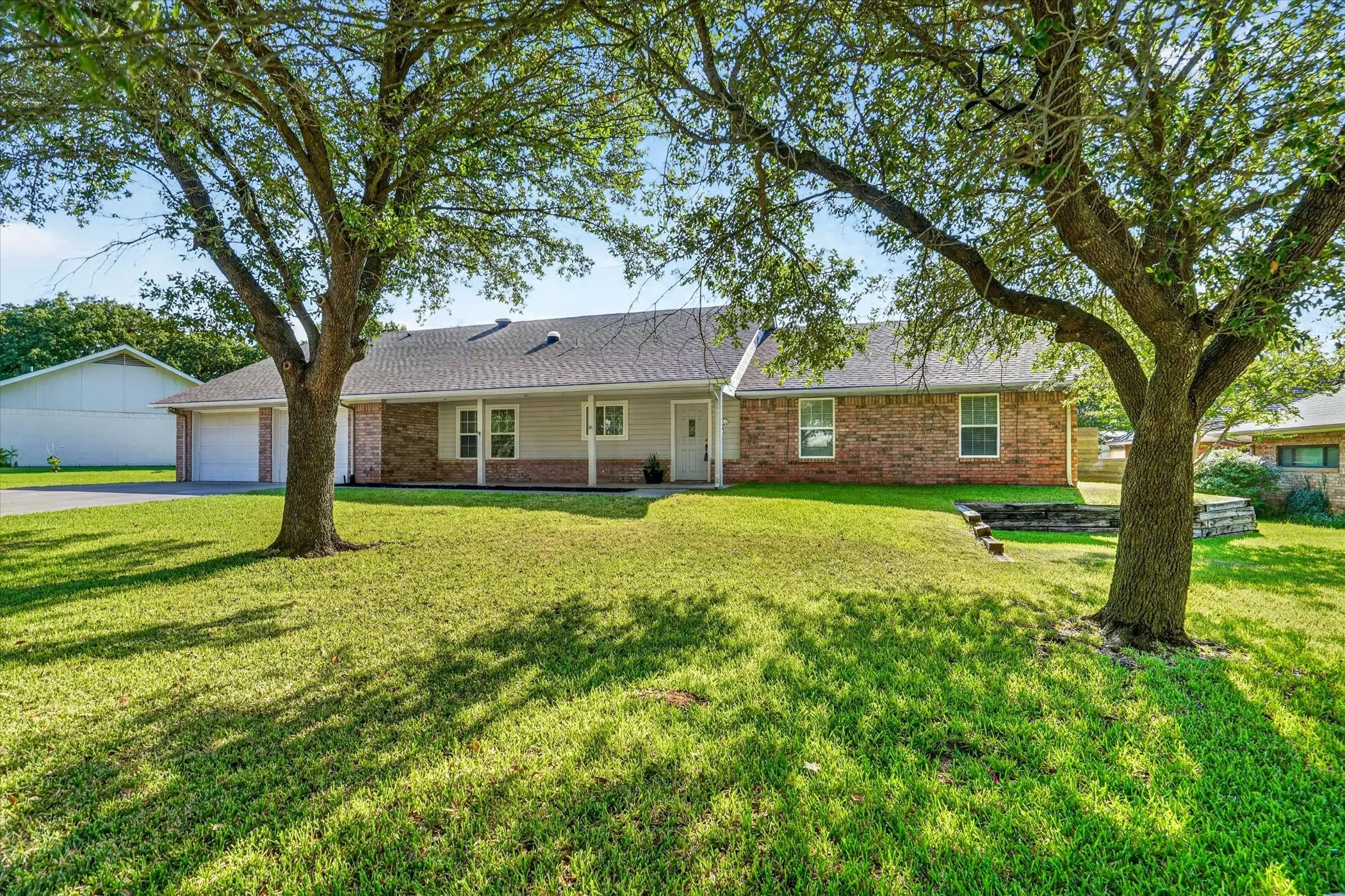 Brick and siding exterior with a covered front porch, two-car garage, mature trees, and a terraced landscape feature