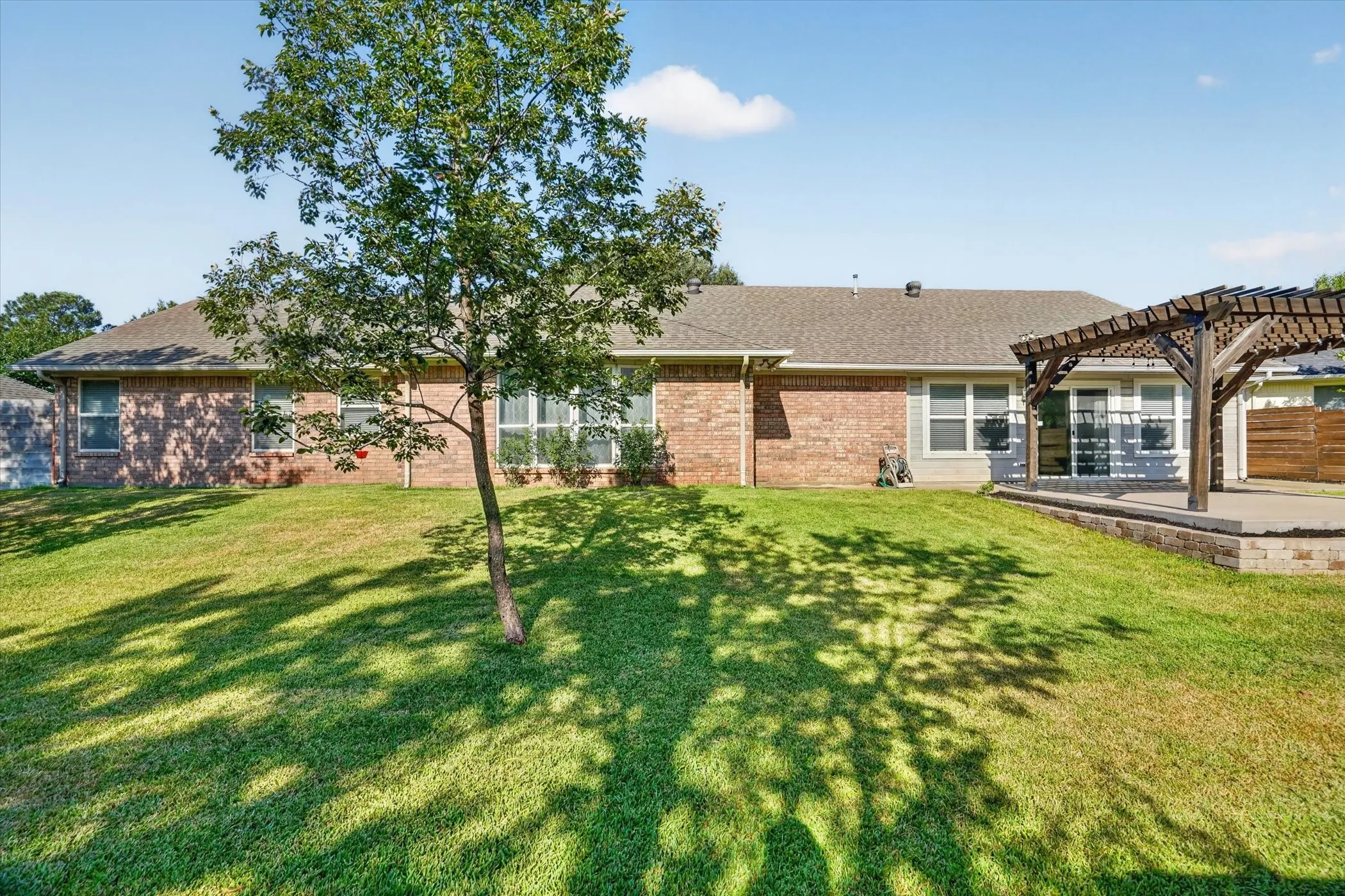 Expansive backyard featuring a mature tree, a brick and siding exterior, multiple windows, a wood pergola, and a raised patio with stone retaining walls