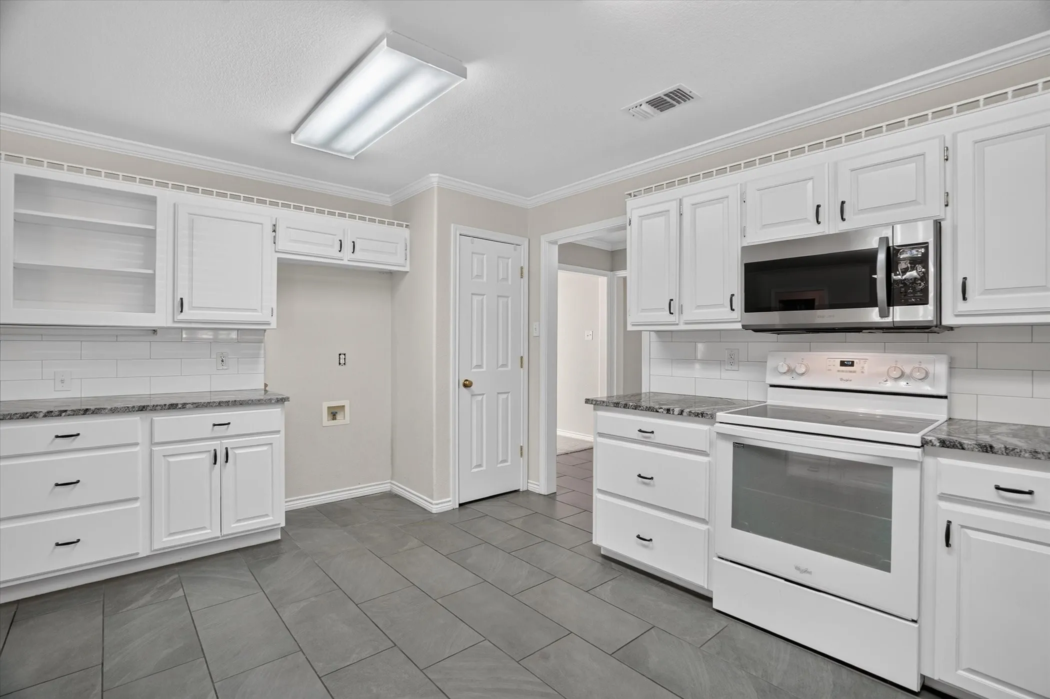 White cabinetry with black hardware, subway tile backsplash, granite-pattern countertops, stainless steel microwave, and tile​​‌​​​​‌​​‌‌​‌‌​​​‌‌​‌​‌​‌​​​‌​​ flooring