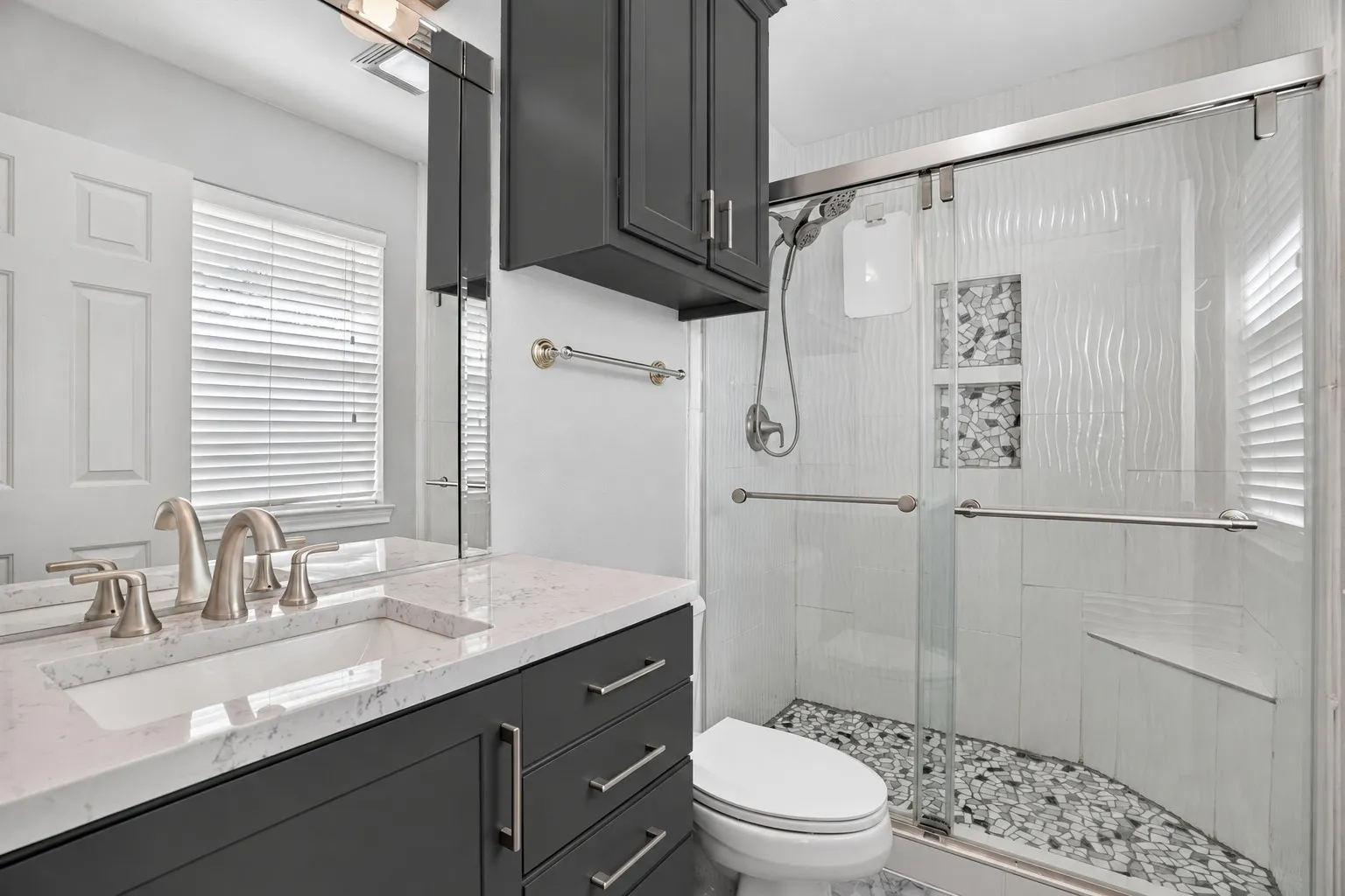 Vanity with a white countertop and undermount sink, dark cabinetry with brushed nickel hardware, and a glass-enclosed shower with a pebble-tiled floor and textured wall tiles