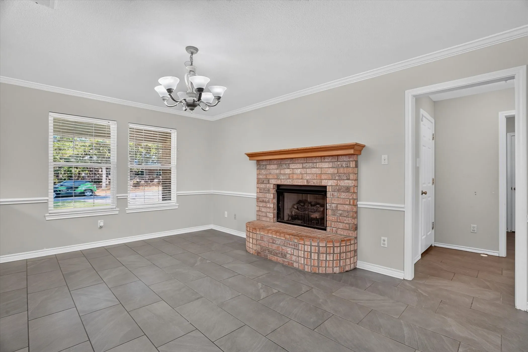 Brick fireplace with wood mantle, tile flooring, two windows, crown molding, and a chandelier
