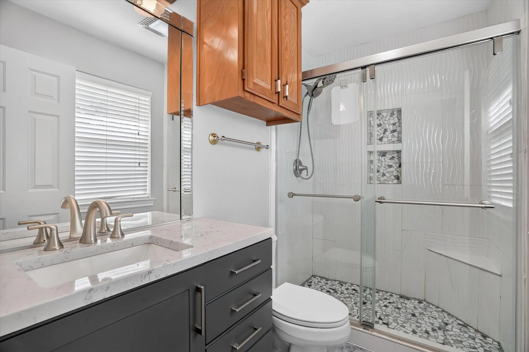 Vanity with a white countertop and undermount sink, dark cabinetry with brushed nickel hardware, and a glass-enclosed shower with a pebble-tiled floor and textured wall tiles