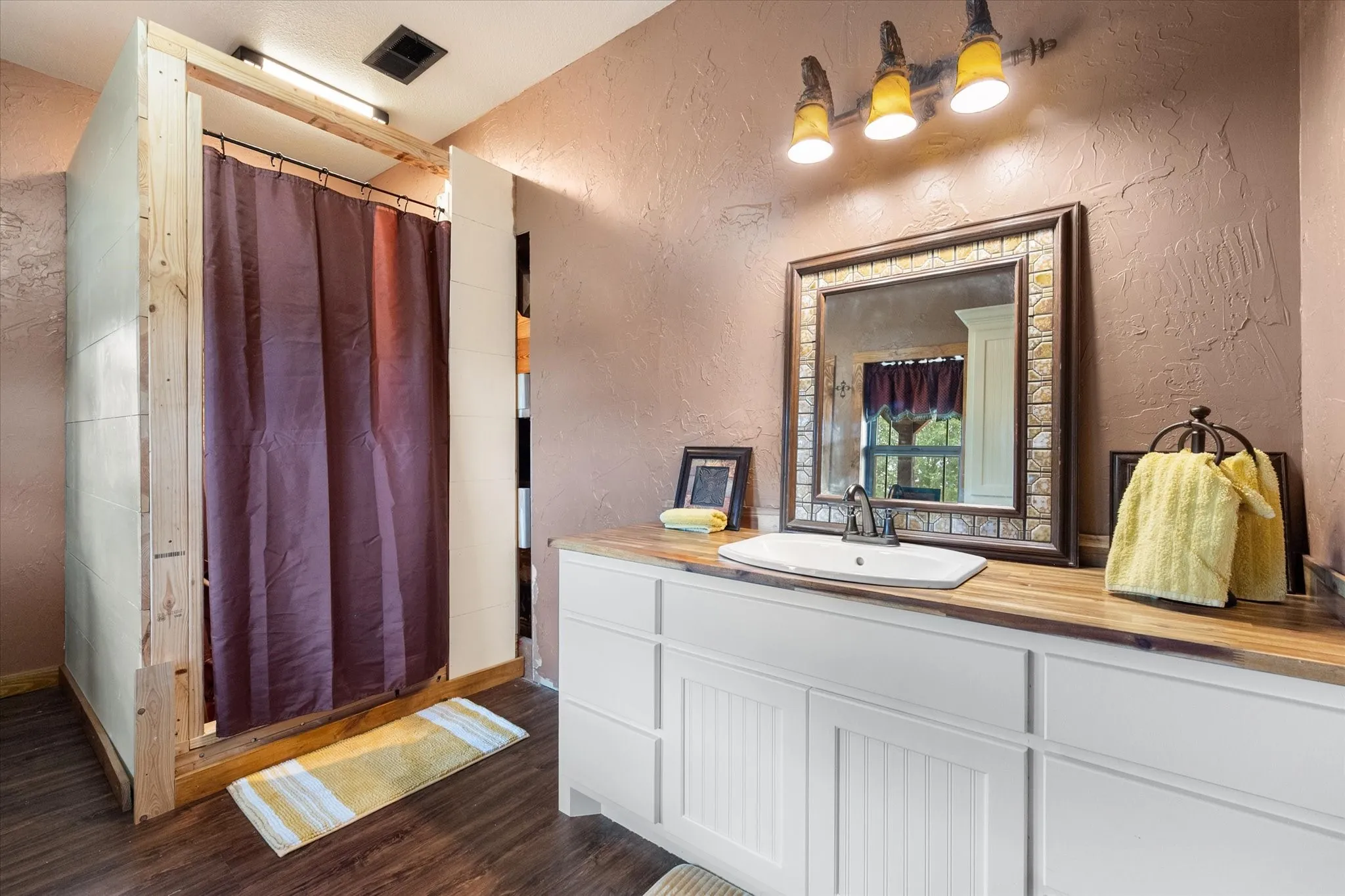 Wood-finish flooring, a freestanding shower enclosure, a vanity with a butcher block-style countertop, an undermount sink, and a decorative framed mirror