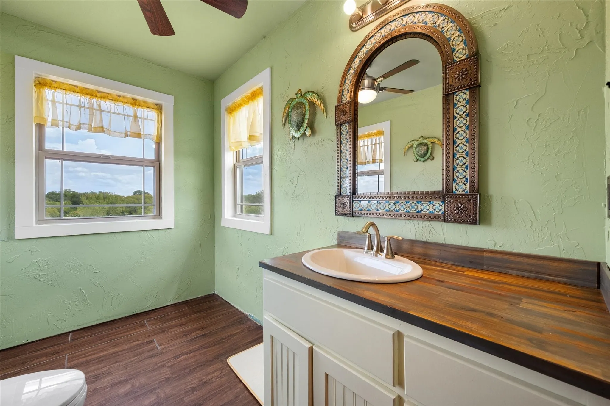 Wood-finish flooring, dual windows, wood countertop vanity with an integrated sink, and an ornate framed mirror with tile accents