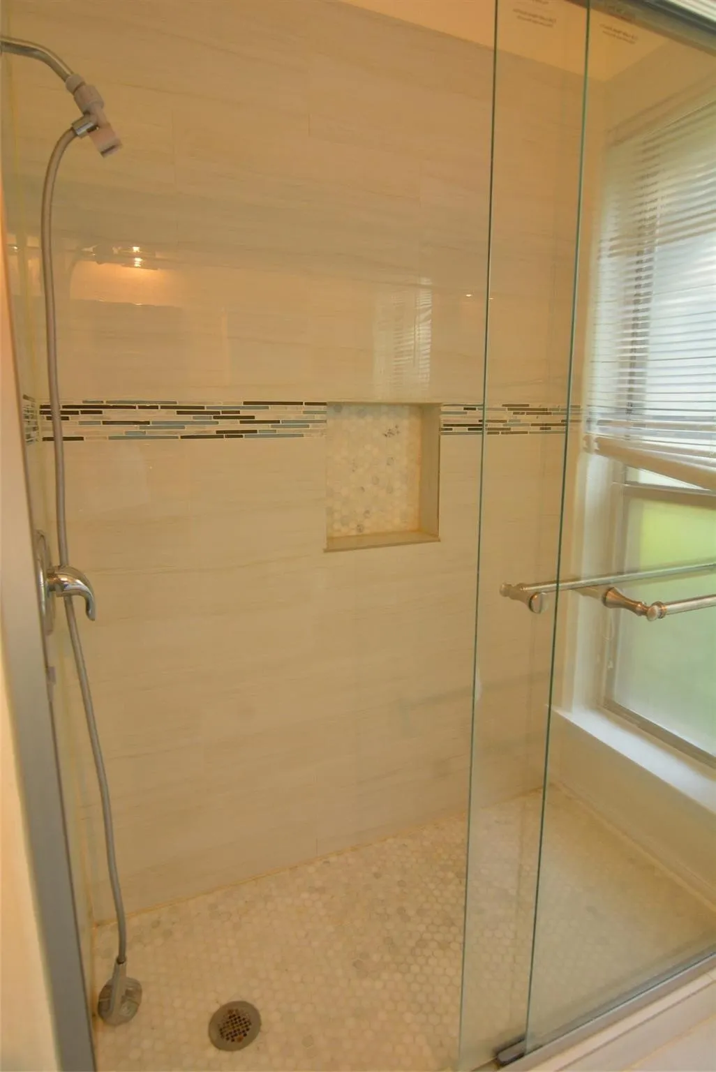 Glass-enclosed shower with a tiled surround, integrated shower niche, mosaic tile accent band, and a window