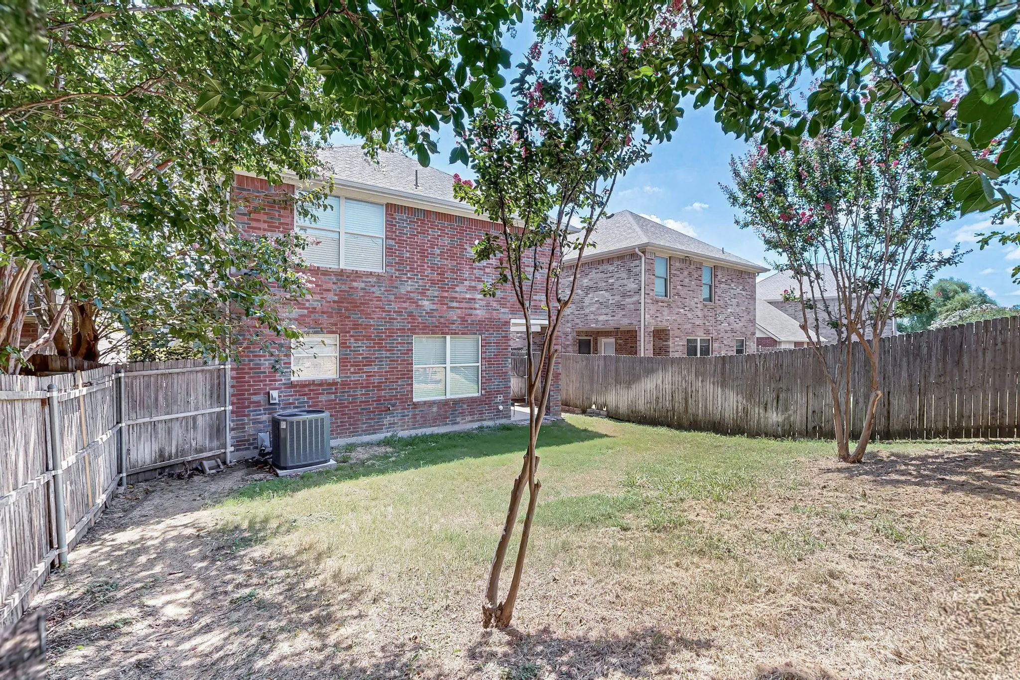Red brick exterior with multiple windows, fenced backyard, mature trees, and central air conditioning unit