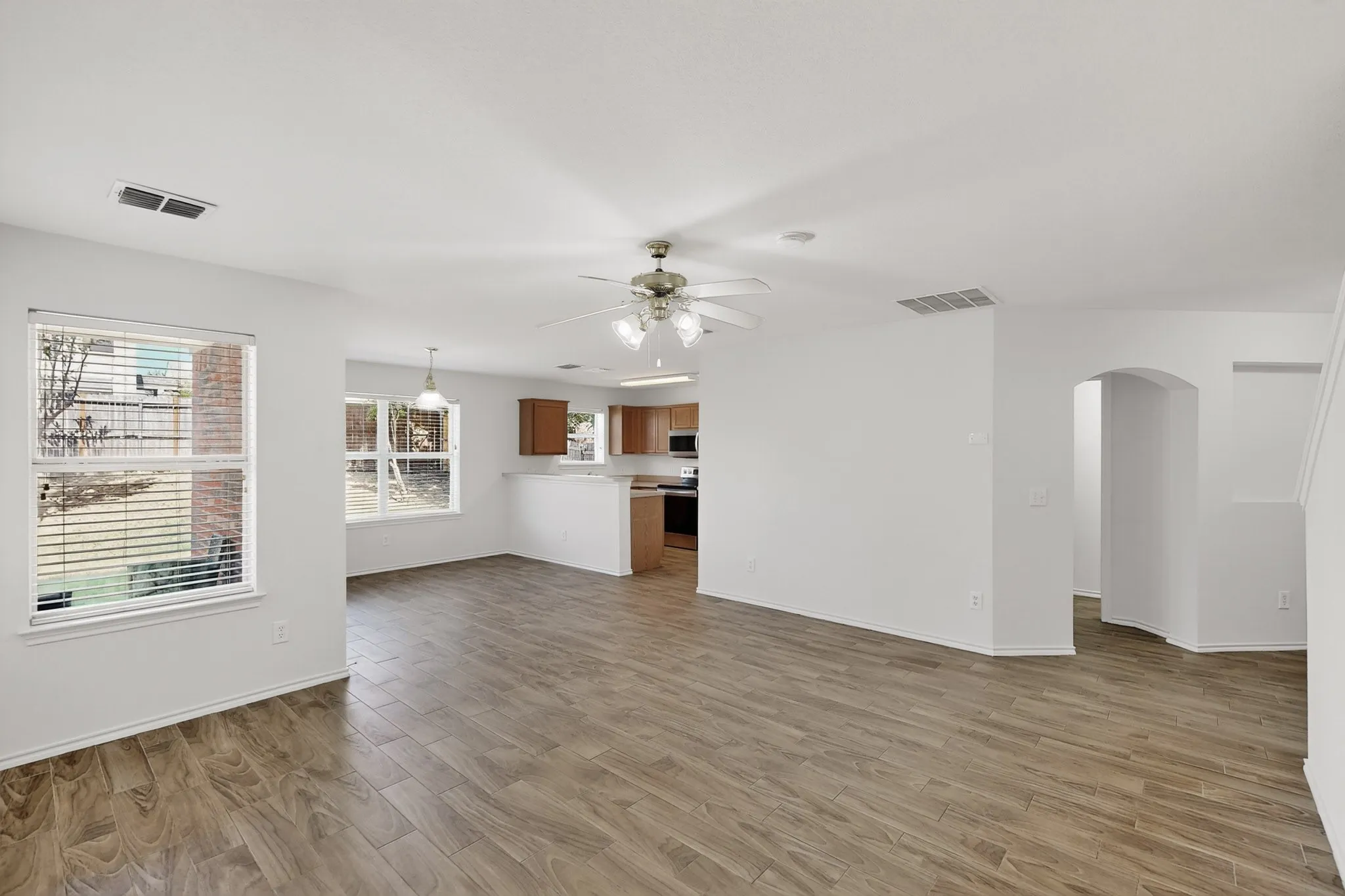 Spacious interior with wood-finish flooring, ample natural light from multiple windows, an arched doorway, and an open-concept kitchen featuring wood cabinetry and a ceiling fan