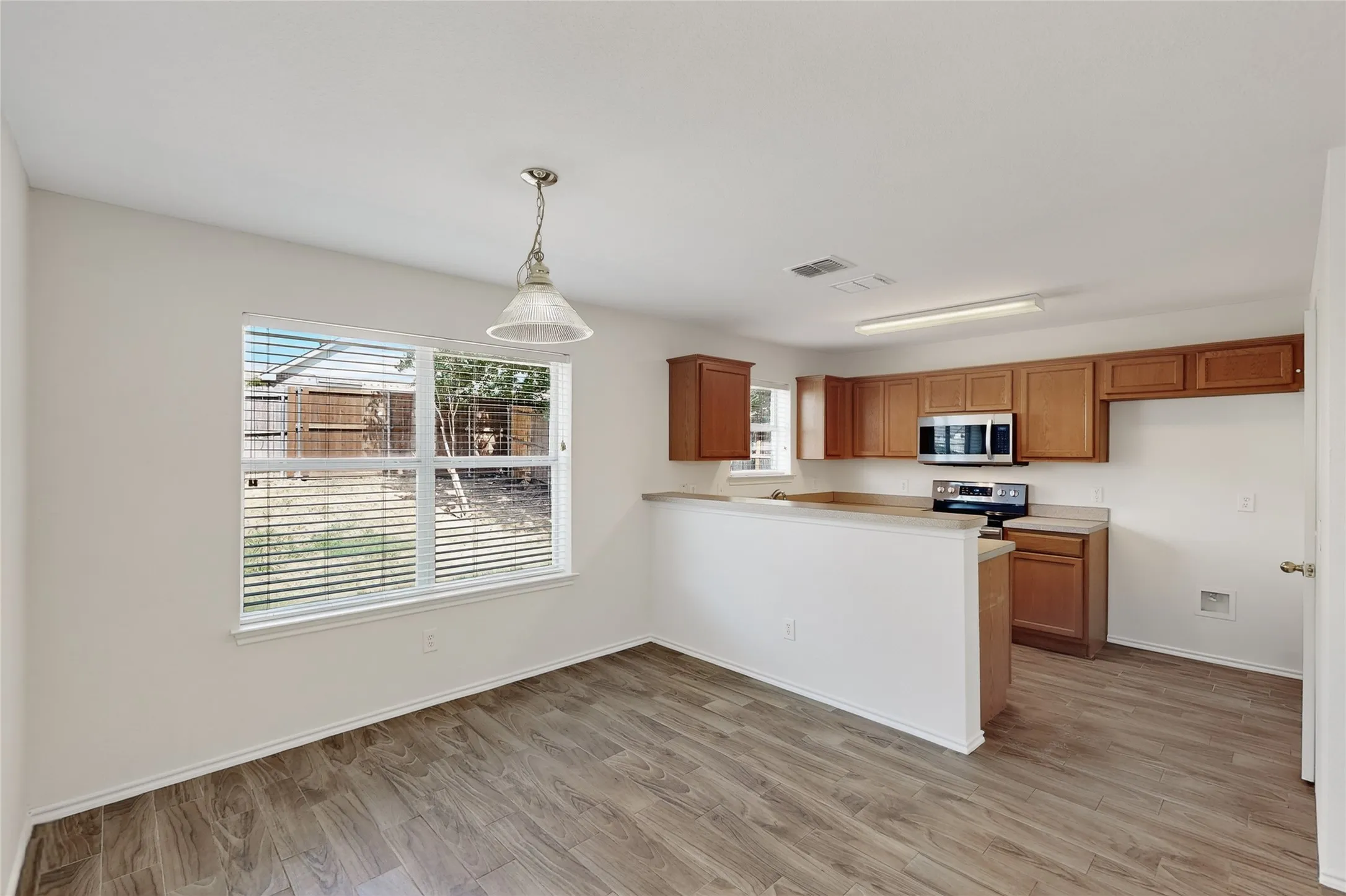 Wood-finish flooring, overhead lighting, kitchen with wood cabinetry, stainless steel appliances, and a window with blinds