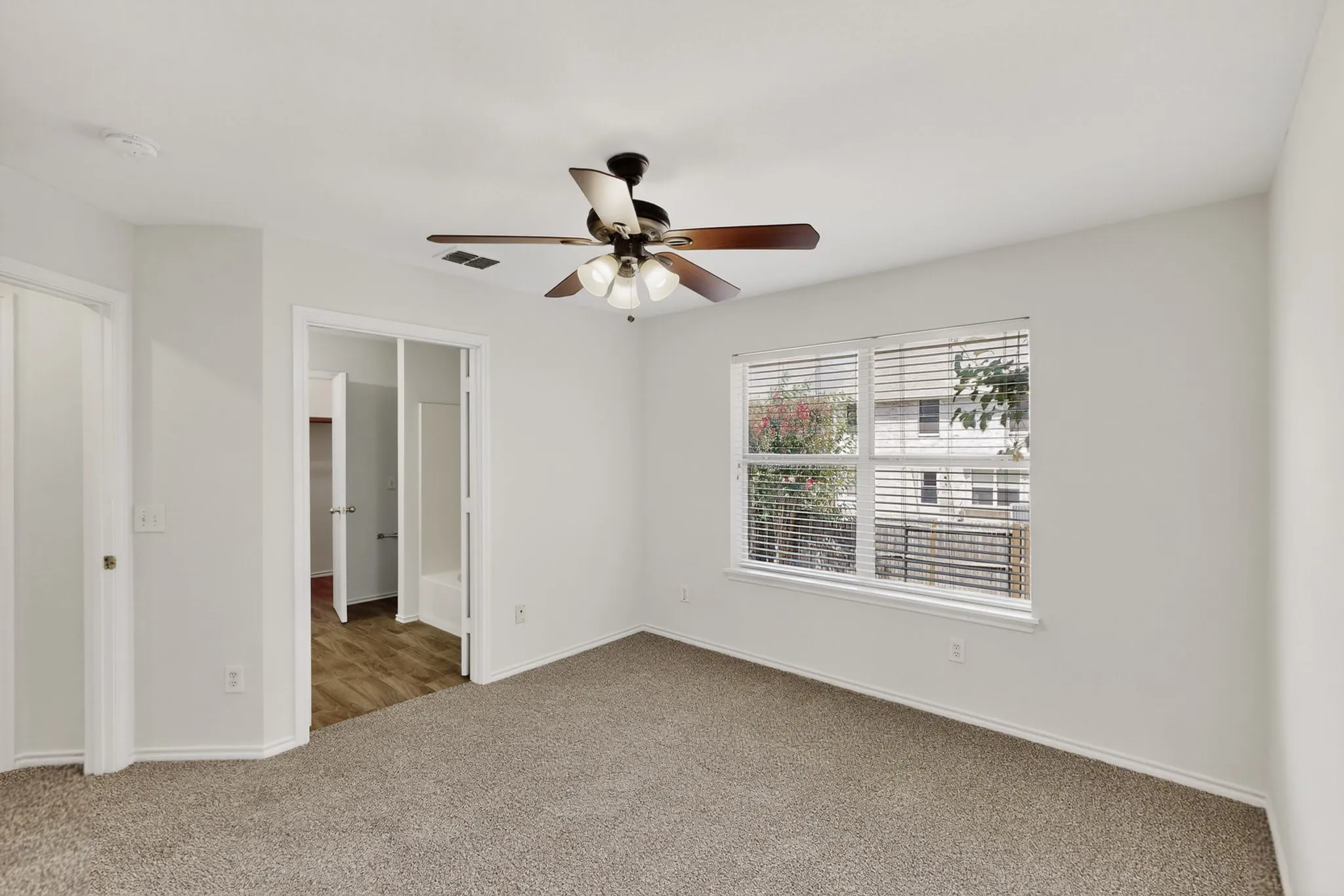 Neutral tone wall paint, textured carpet flooring, ceiling fan with light fixture, window with horizontal blinds, and wood-finish flooring in an adjoining room