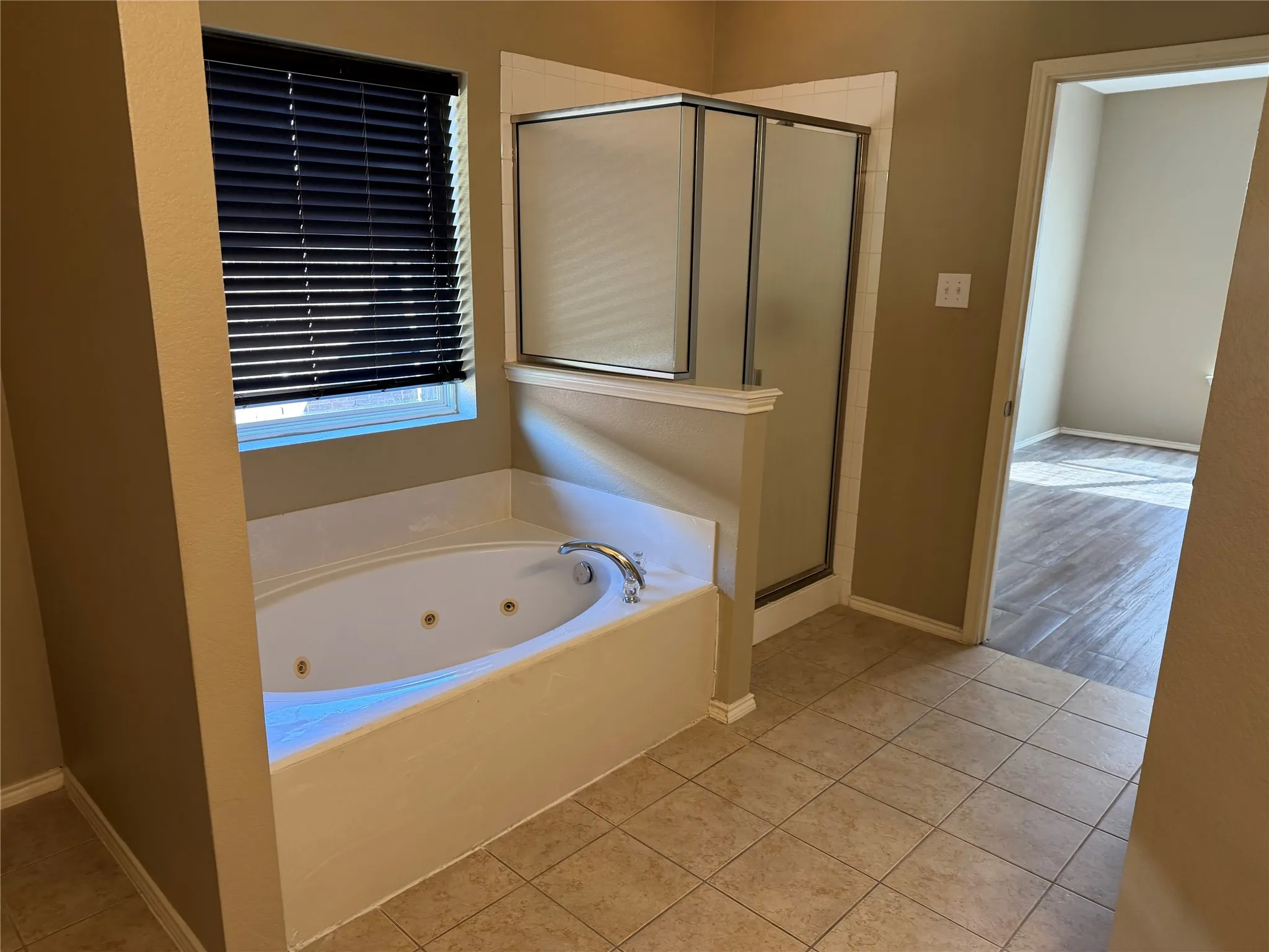 Soaking tub with integrated jets, glass-enclosed shower, tiled flooring, and a window with blinds