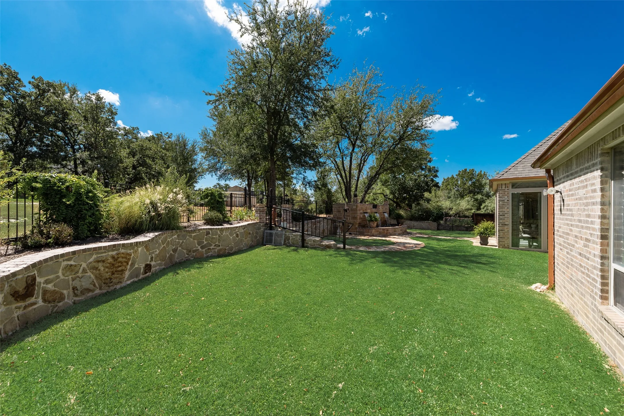 Expansive lawn area, stone retaining wall with integrated landscaping, mature trees, brick exterior, and covered patio access