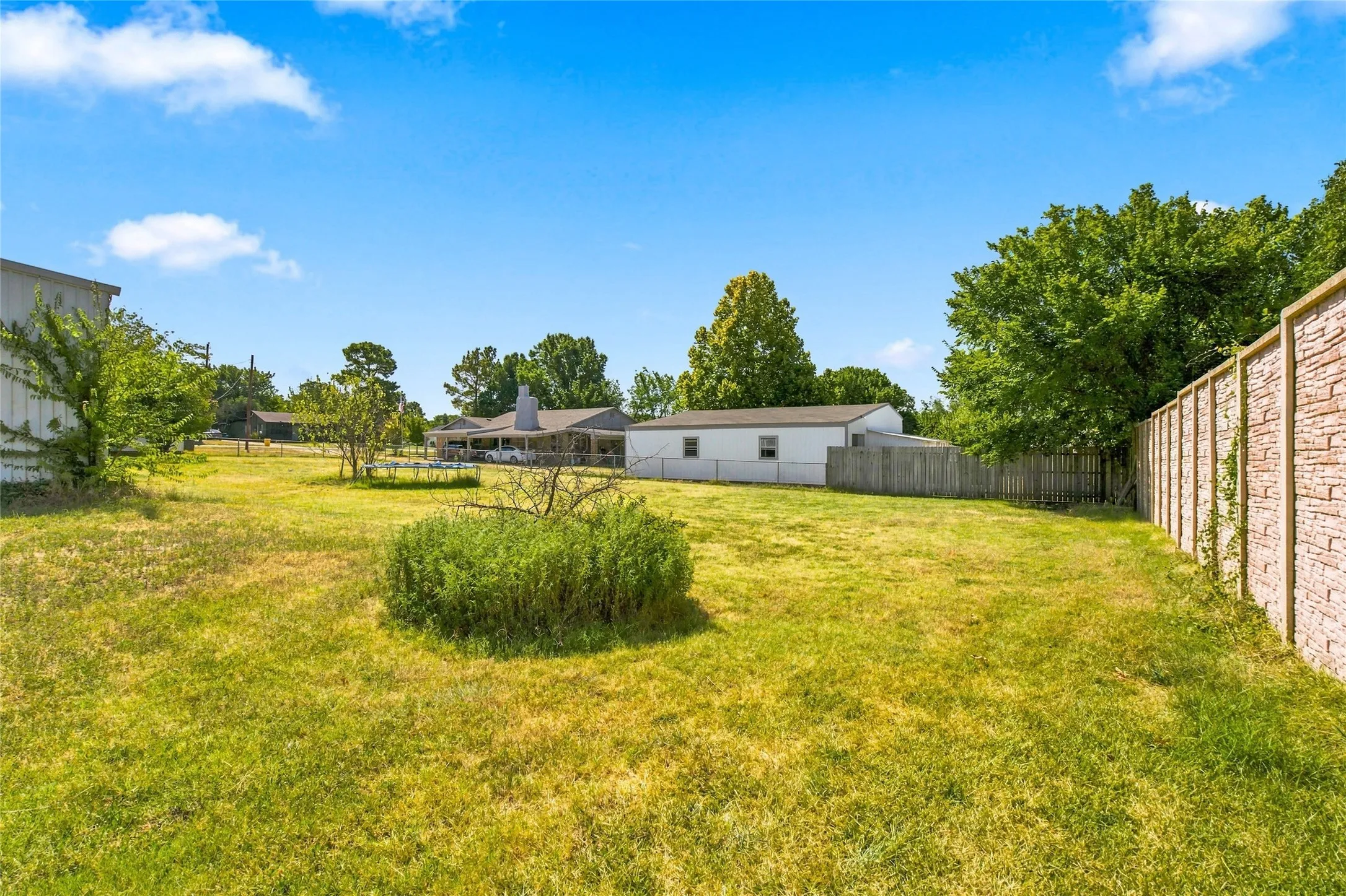 Expansive grassy lawn with established trees, featuring a decorative stone-finish privacy wall and a white wood-panel fence