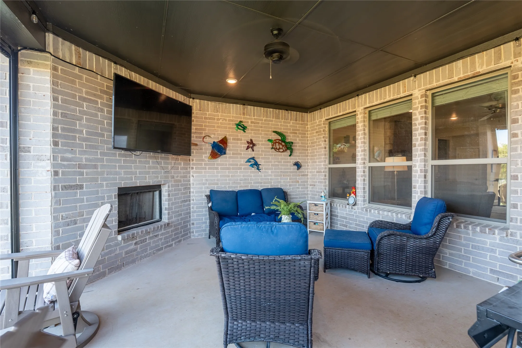 Covered outdoor patio featuring a brick fireplace, built-in television alcove, ceiling fan, and multiple large windows