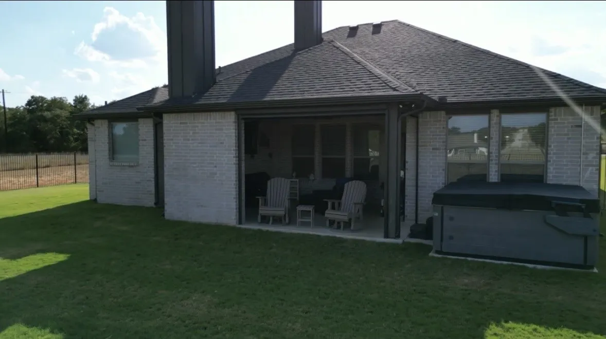 Brick exterior with a covered patio, shingled roof, and an outdoor spa