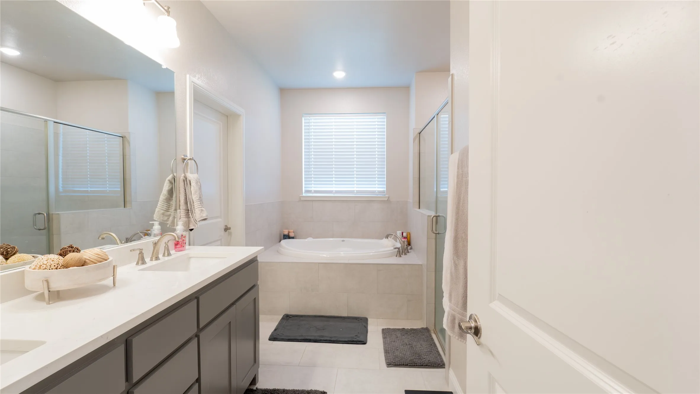 Recessed lighting, light-toned tile flooring, built-in bathtub, dual-sink vanity with light-toned countertop, and a glass-enclosed shower