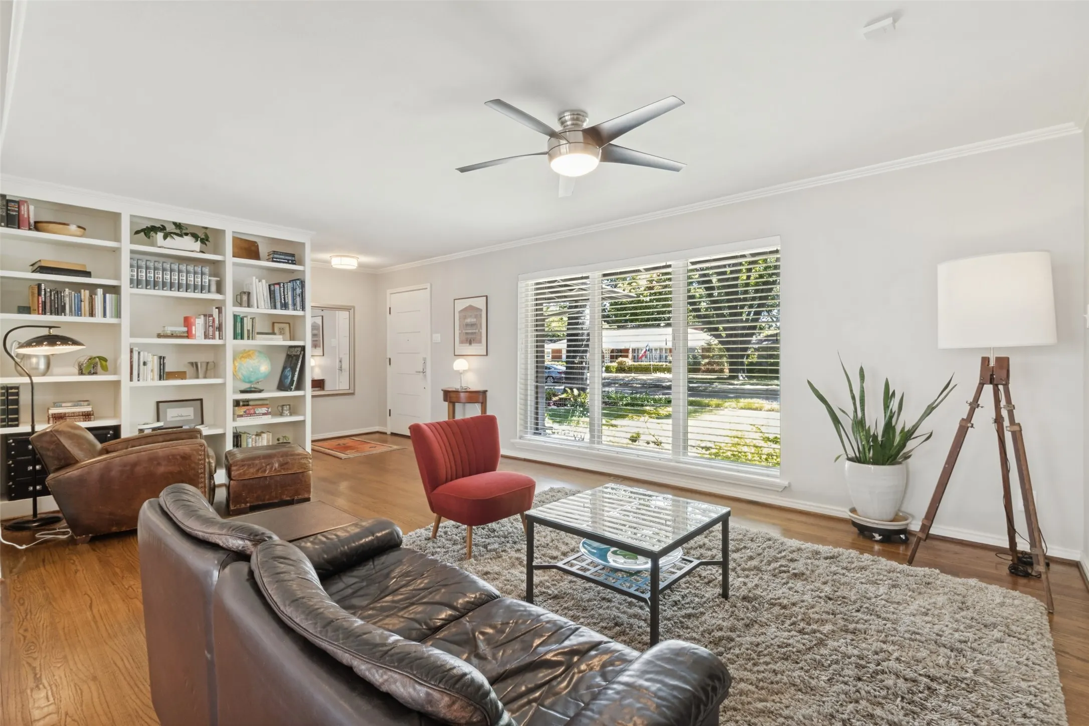Original Hardwood floors, crown molding, white built-in shelving, large windows with blinds, and a ceiling fan with integrated lighting