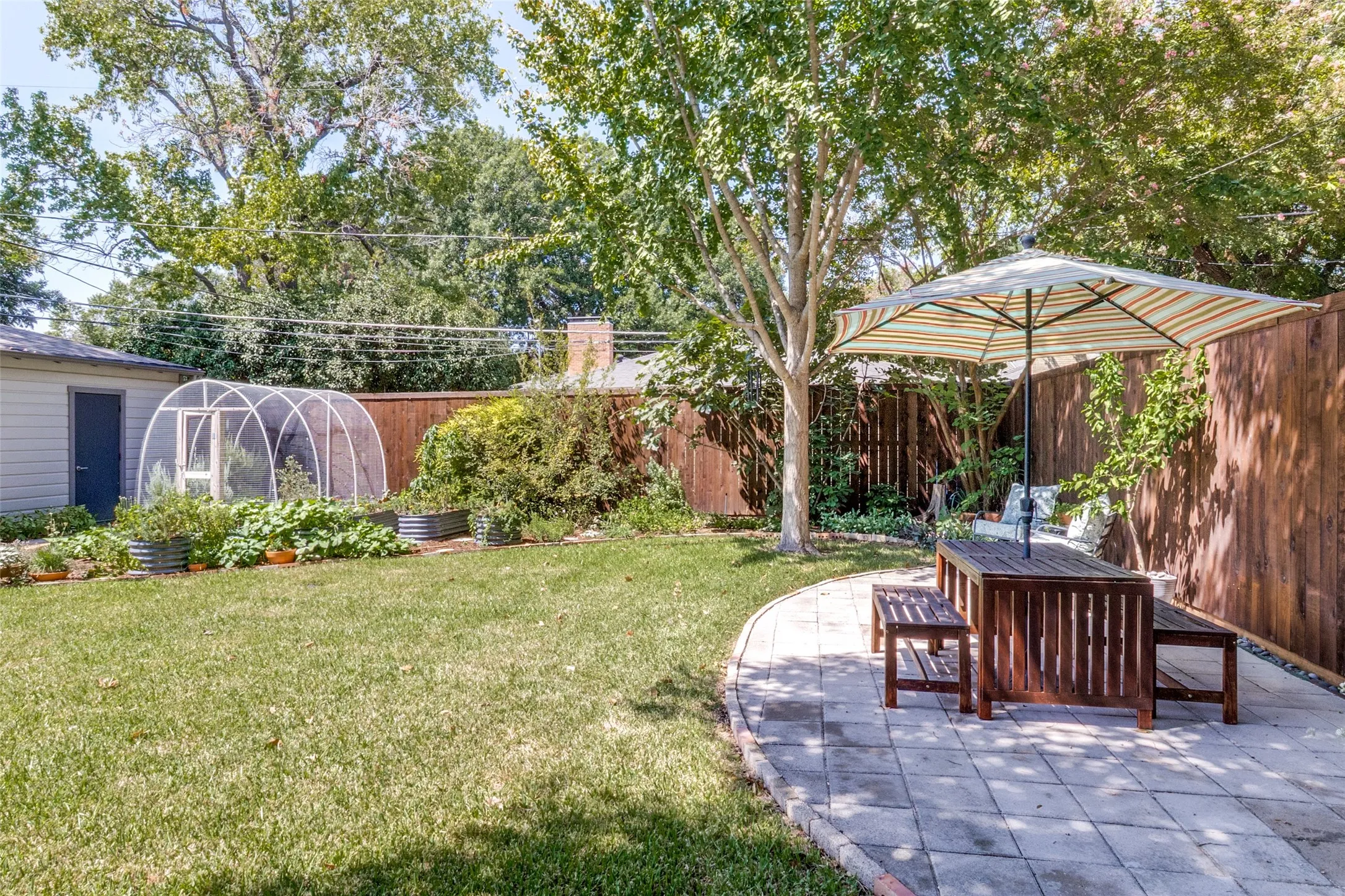 Expansive backyard featuring a manicured lawn, established shade trees, a curved paver patio, and a dedicated greenhouse structure