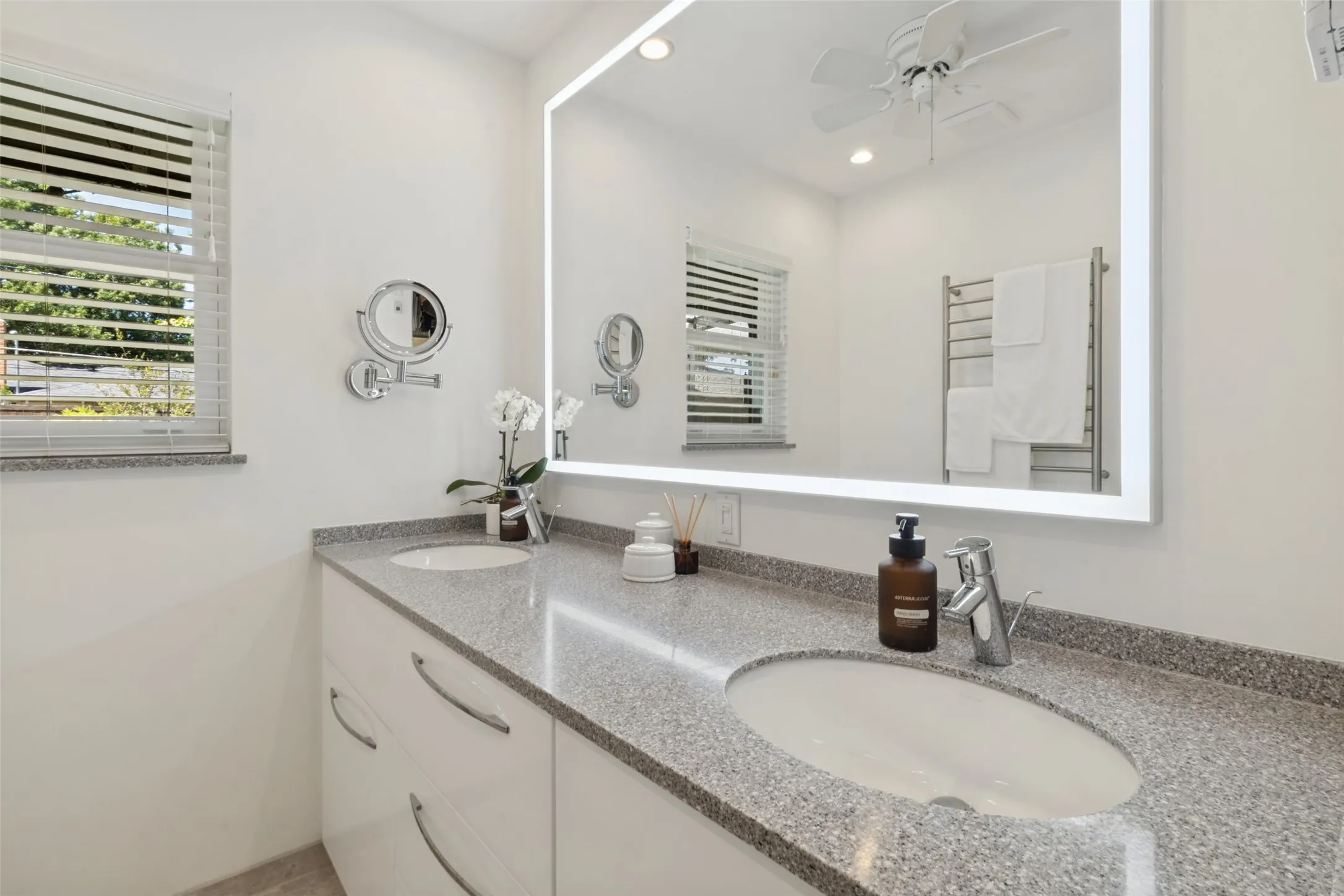 Dual undermount sinks with chrome fixtures, a spacious countertop, an illuminated vanity mirror, and a wall-mounted towel warmer
