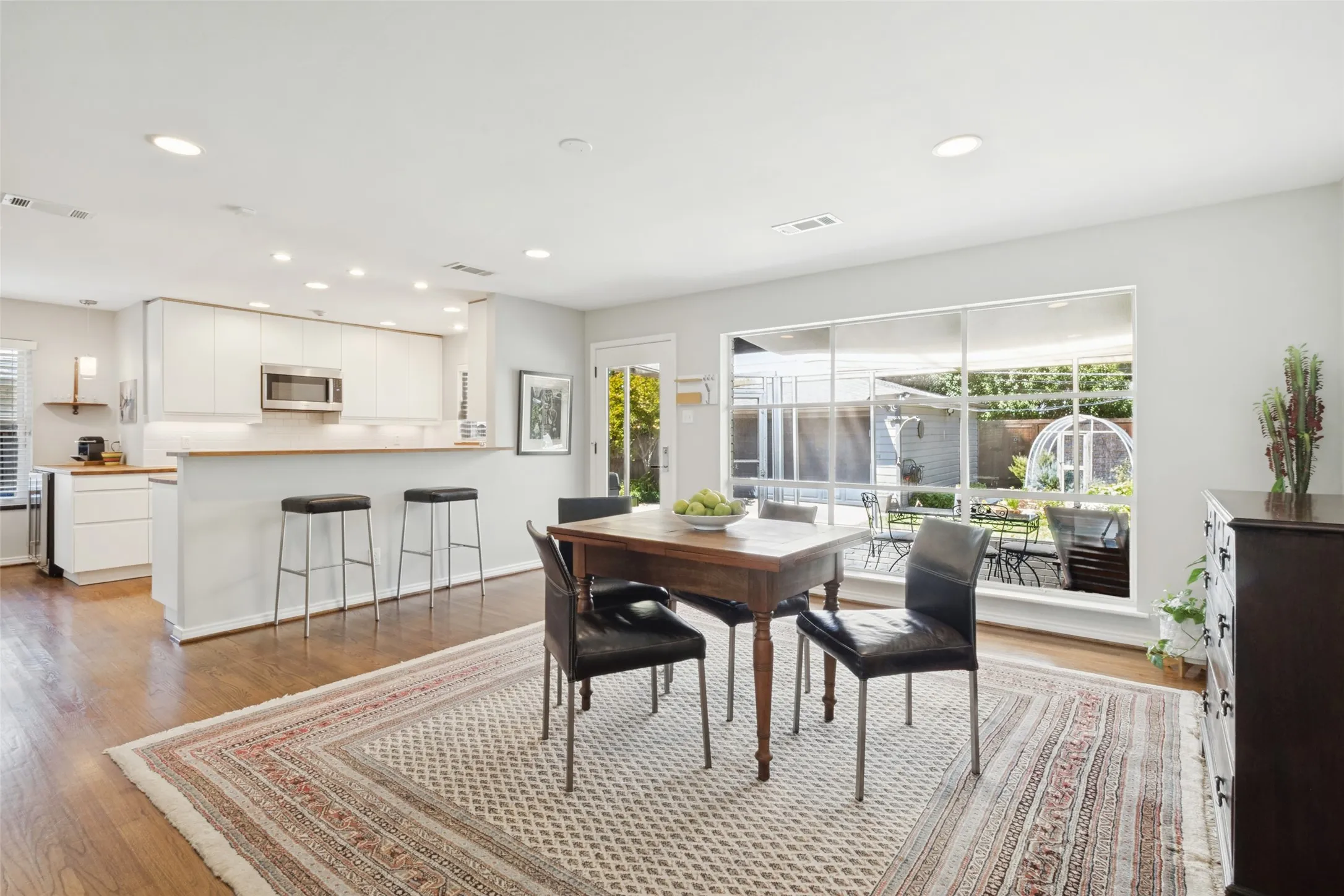 Wood-finish flooring throughout, kitchen with white cabinetry and stainless steel microwave, breakfast bar with pendant lighting, recessed ceiling lighting, and large windows overlooking a private outdoor space