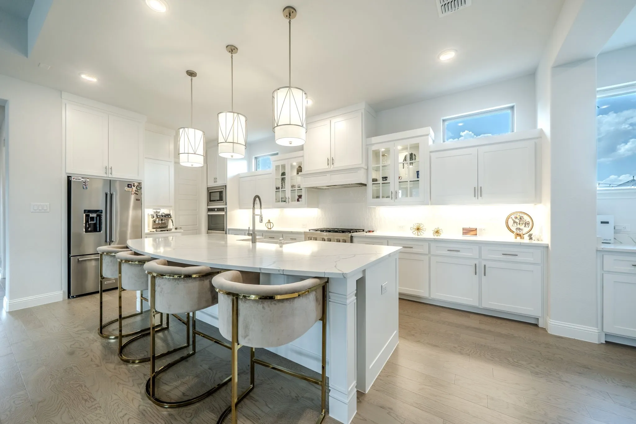 Spacious kitchen featuring an extended island with a built-in sink, white shaker cabinetry, stainless steel appliances, pendant lighting, and wood-finish flooring