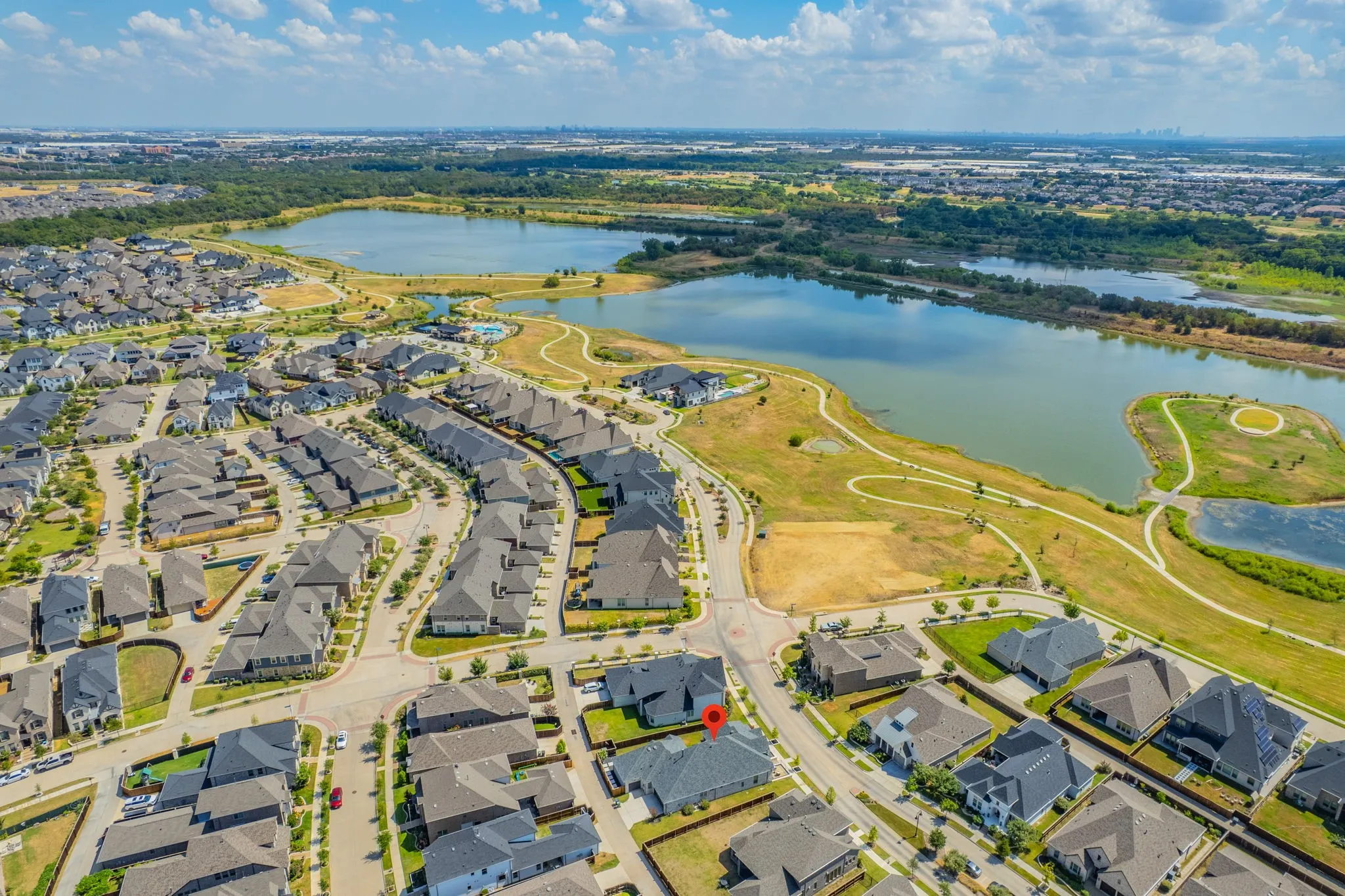 Aerial perspective of a developed residential community featuring numerous single-family homes with dark-toned roofs, streets with planted trees, and a prominent lake with walking paths